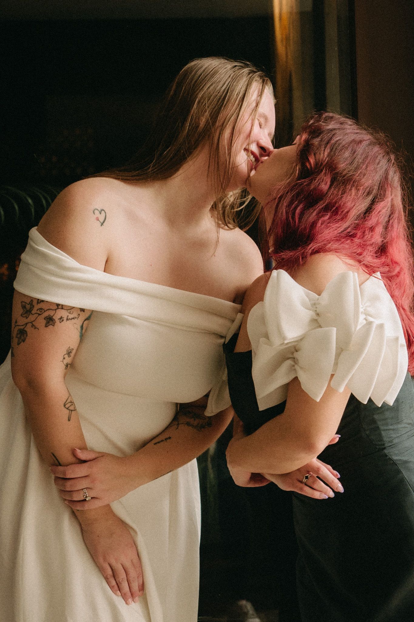 Two brides sharing a kiss, one with tattoos in off-shoulder gown, other in white bow dress.