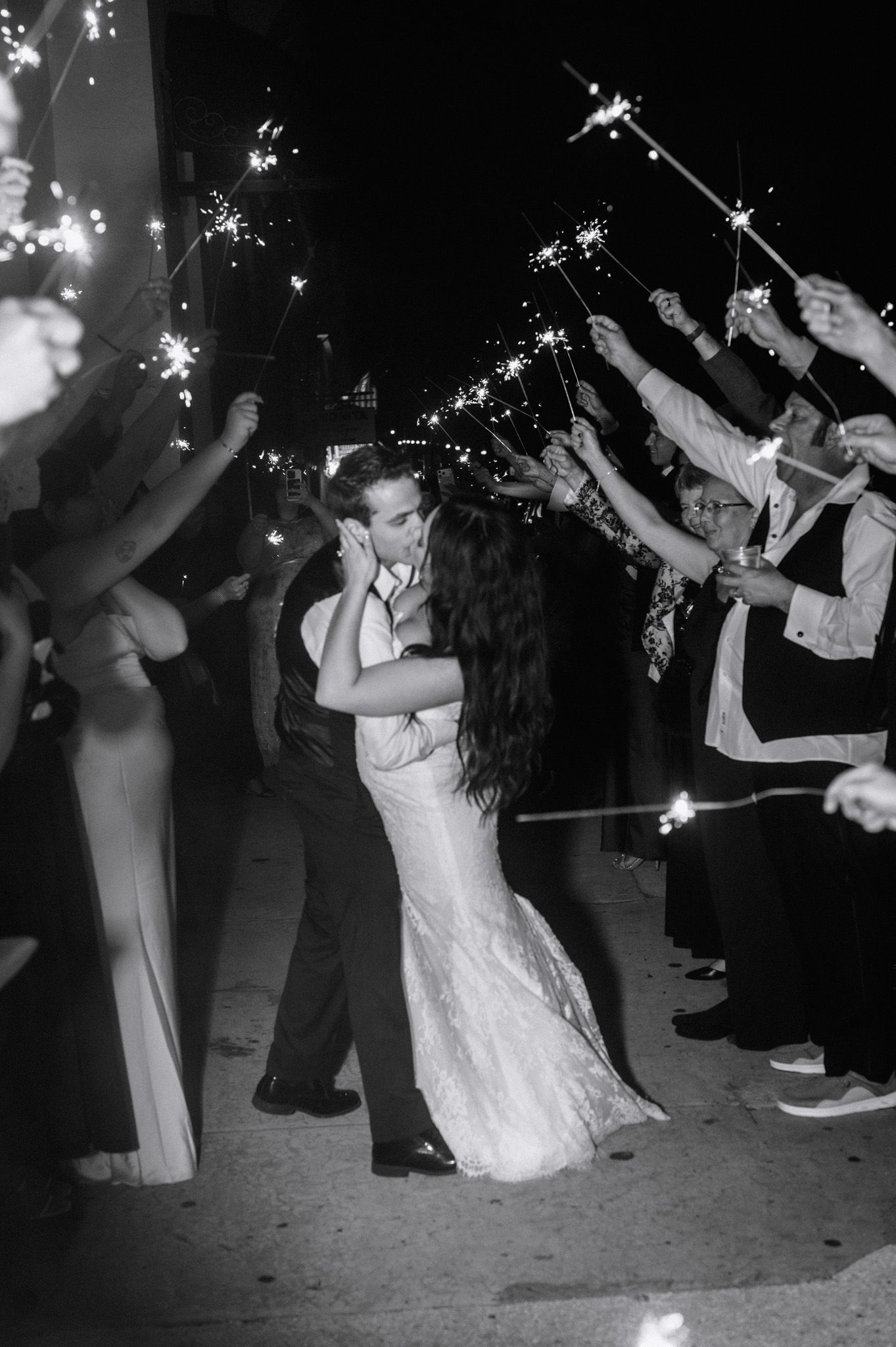Black and white photo of newlyweds kissing under sparklers held by guests during wedding send-off.