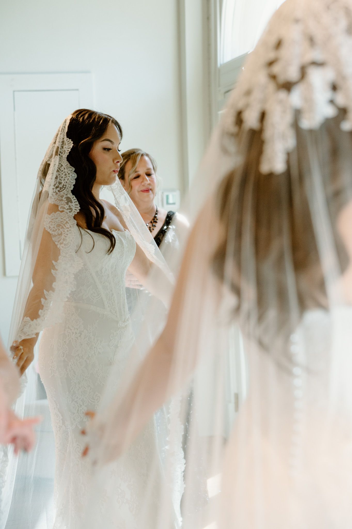 Bride in lace wedding gown and veil being assisted by older woman, with mirror reflection visible in bright room.