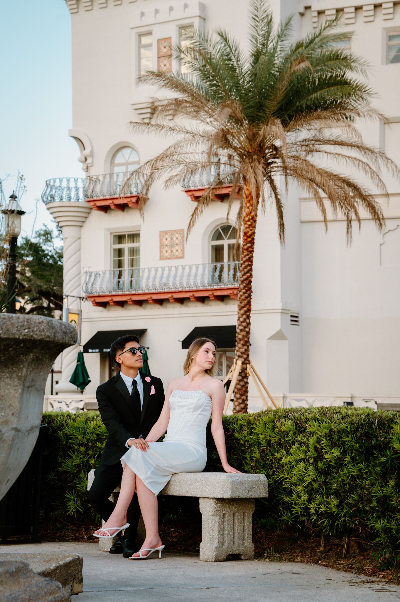 Bride and groom posing on stone bench beside palm tree in front of elegant white building.