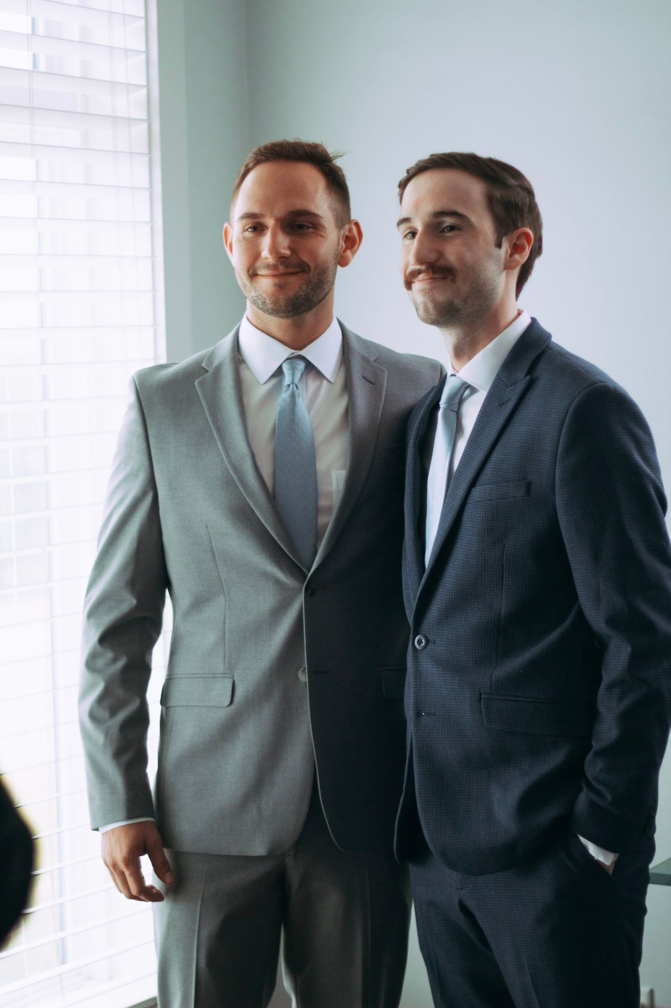 Two smiling groomsmen posing together in gray and navy blue suits near bright window.