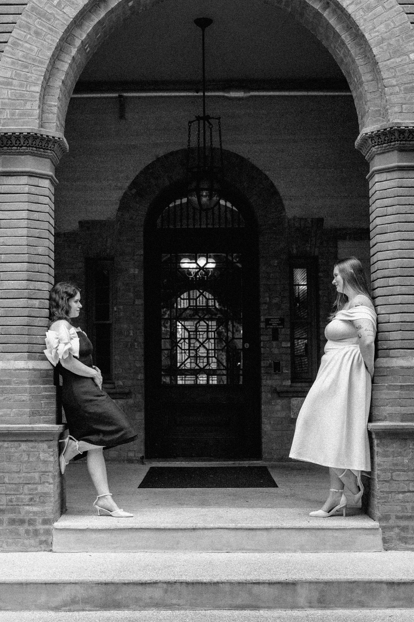 Two women in elegant dresses posing under historic brick archway in black and white photo.