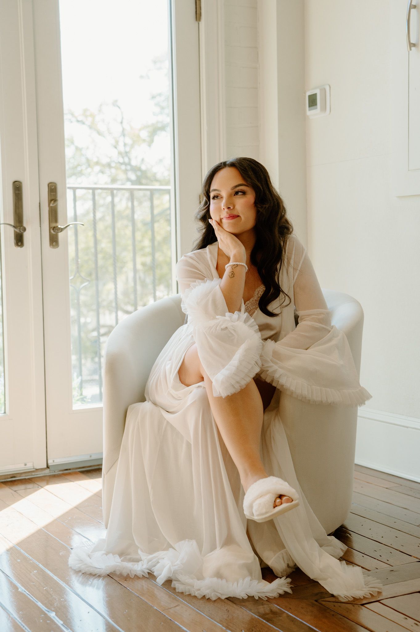 Bride in white tulle robe and feather slippers sits gracefully in armchair by sunlit French doors.