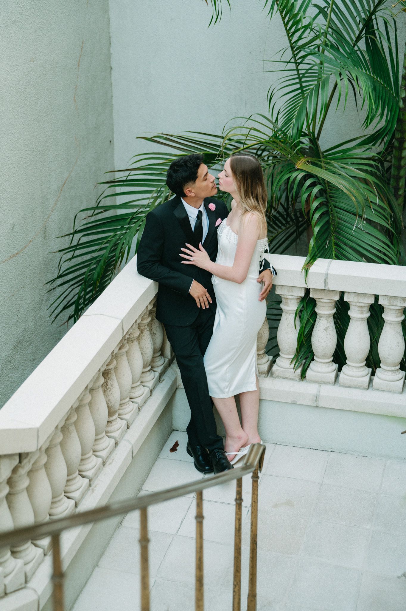 Couple sharing a kiss on a balcony with tropical palm trees and white balustrade.
