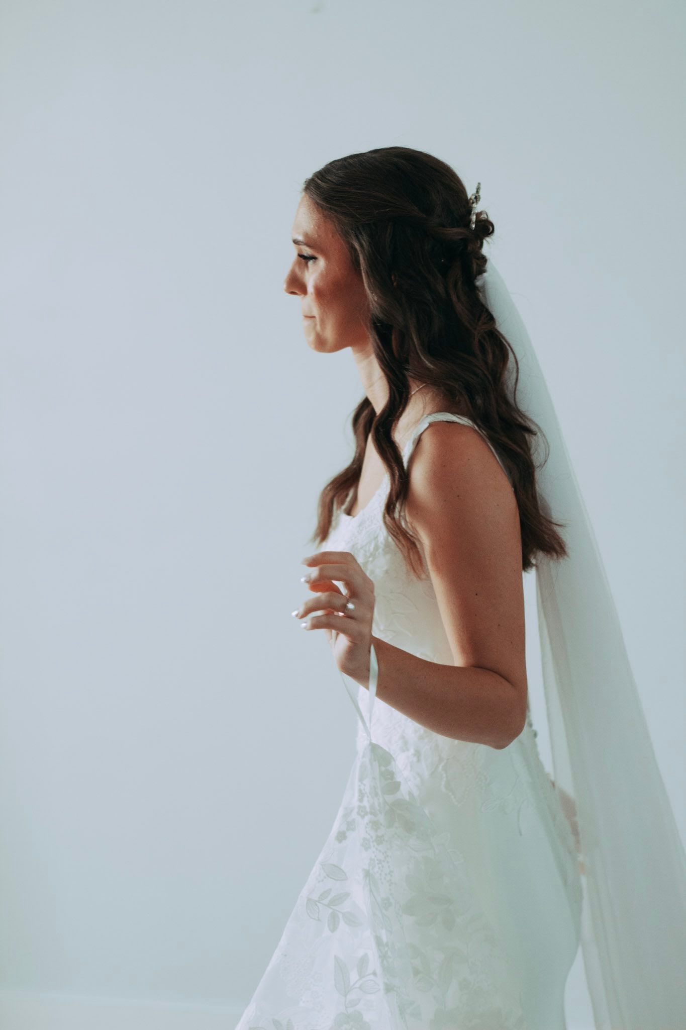 Beautiful bride in white lace gown with veil posing in profile against soft white background.