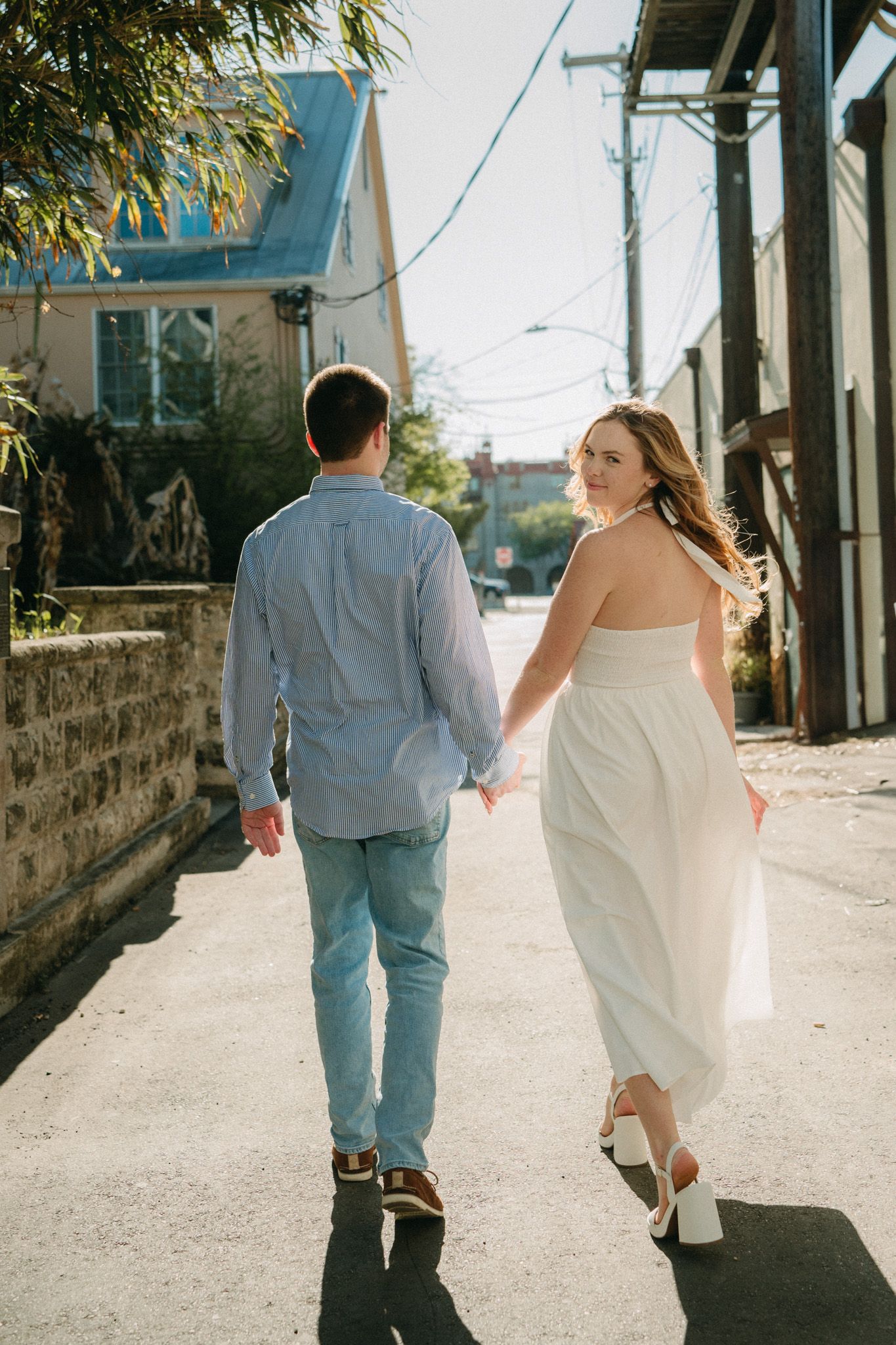 Couple walking hand-in-hand down a sunny alley, woman in white strapless dress glancing back, man in blue shirt and jeans.