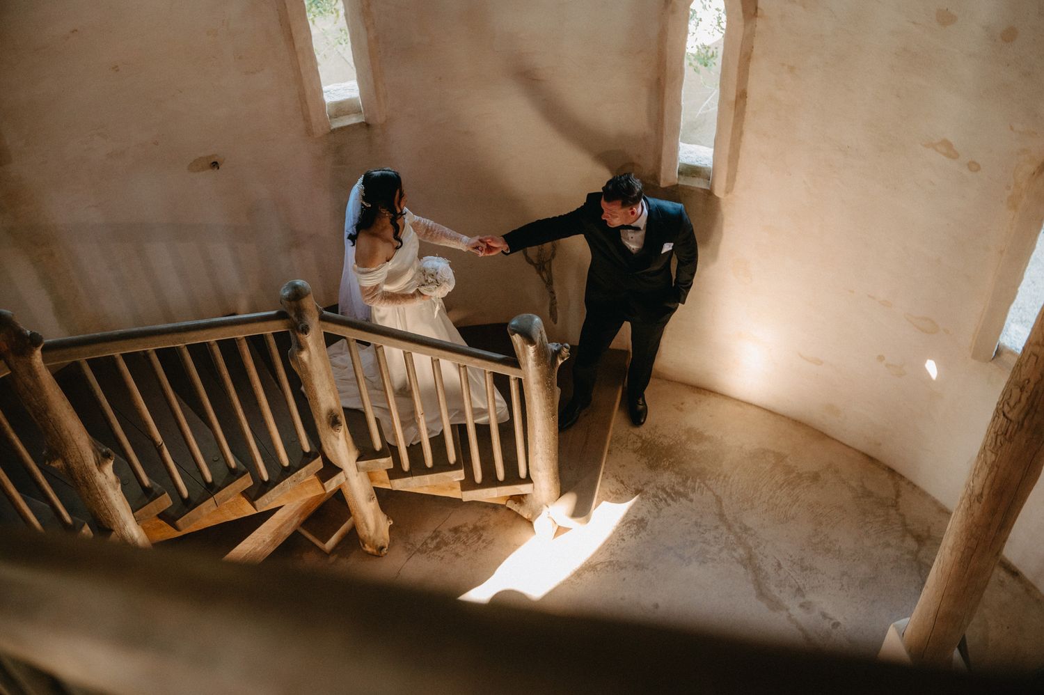 Bride and groom hold hands on rustic wooden spiral staircase in a warm-toned venue during wedding celebration.
