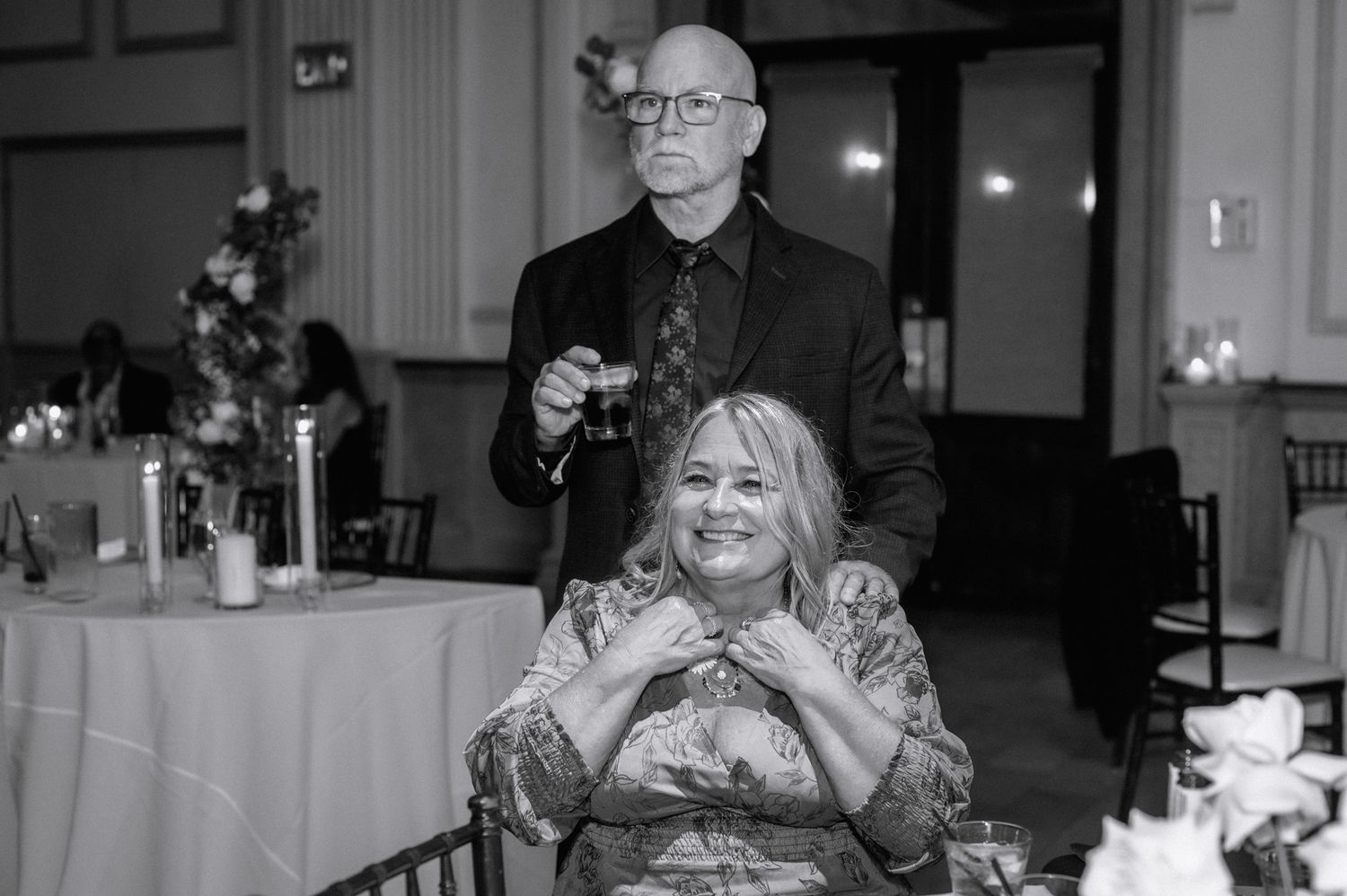 Black and white photo of a smiling woman and bald man in formal attire at an elegant wedding reception.