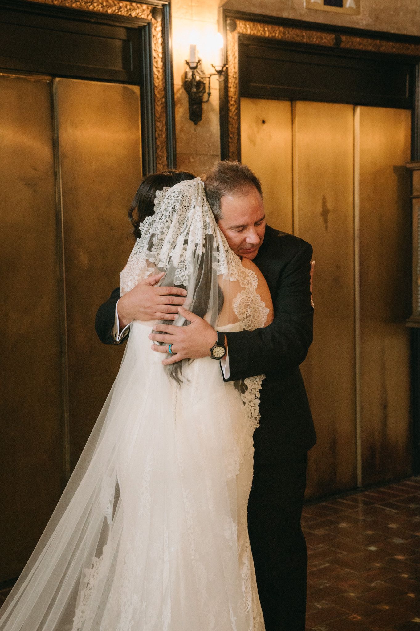 Bride in lace wedding gown and veil sharing an emotional hug with a man in a black suit at a wedding venue.