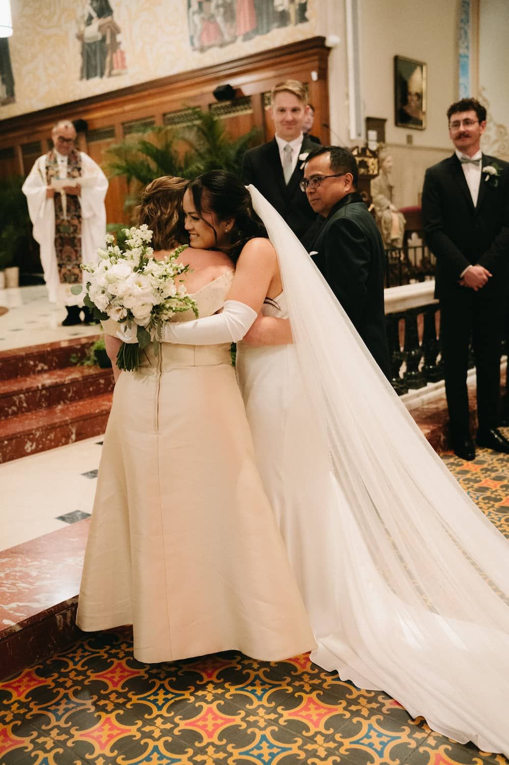 Bride hugging a family member at the altar during a church wedding ceremony, holding a white floral bouquet.