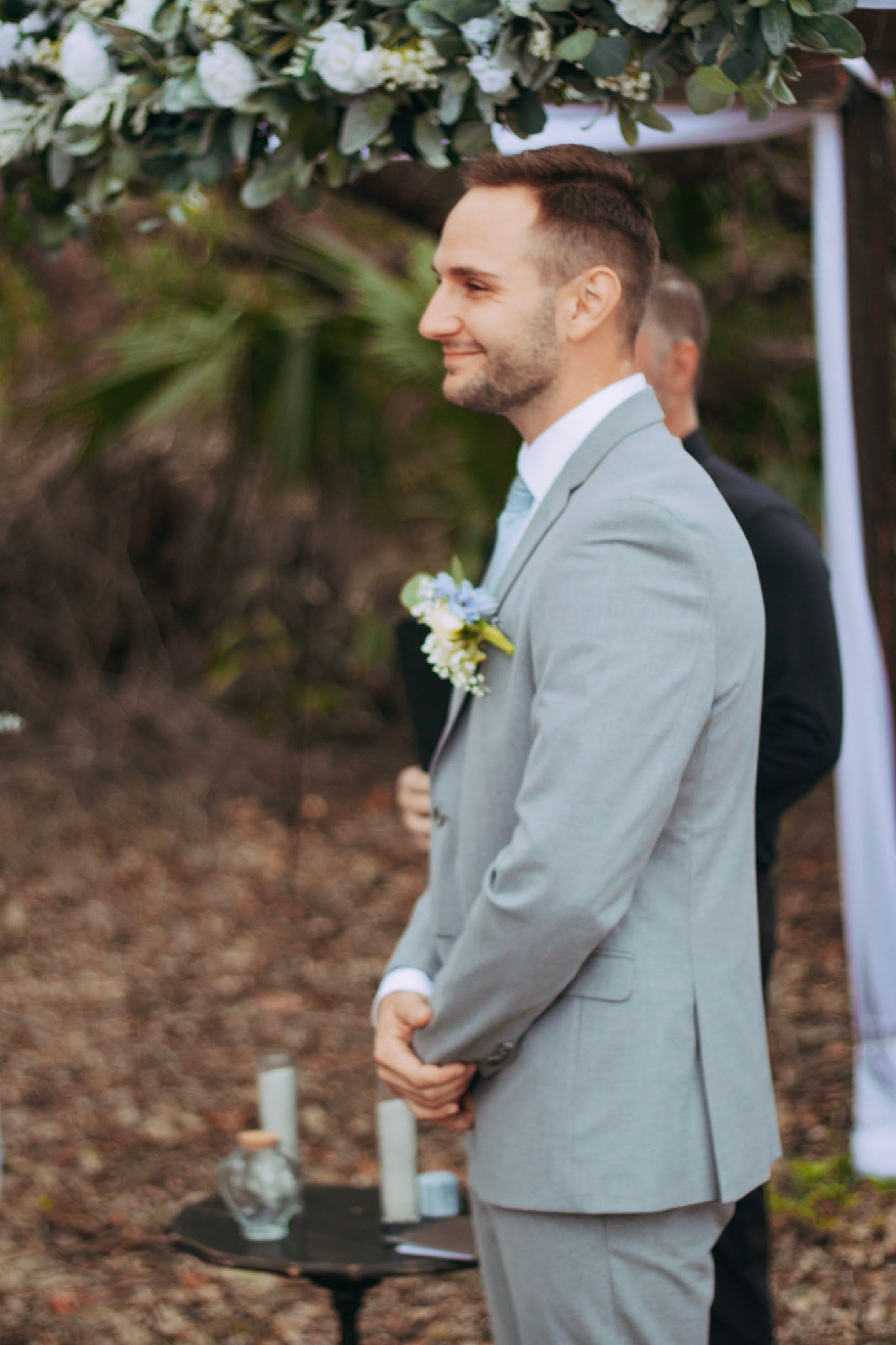 Smiling groom in light gray suit with boutonniere stands at outdoor wedding ceremony altar.