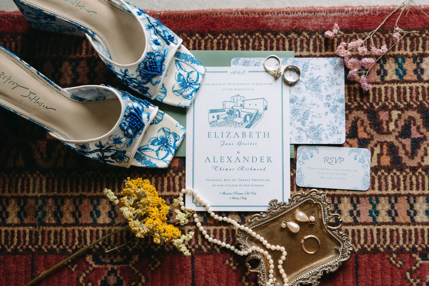 Blue floral wedding shoes beside elegant invitation suite, gold rings, pearl jewelry, and dried flowers on a red patterned rug.