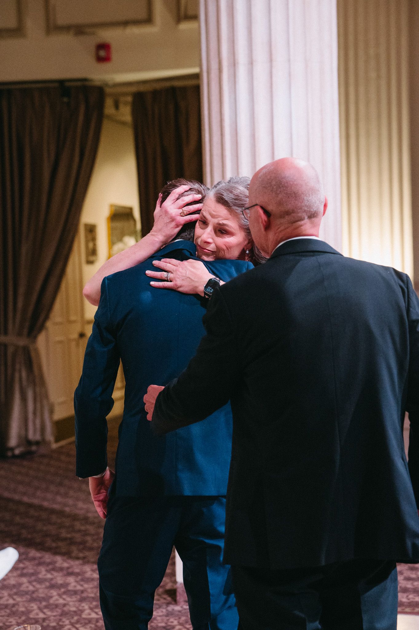Emotional wedding moment as an older woman embraces a man in a blue suit while a bald man looks on.