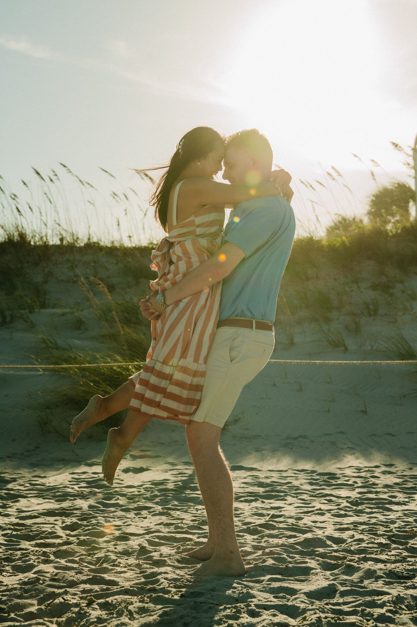 Man lifting woman in striped dress on sandy beach with golden sunset backlight.
