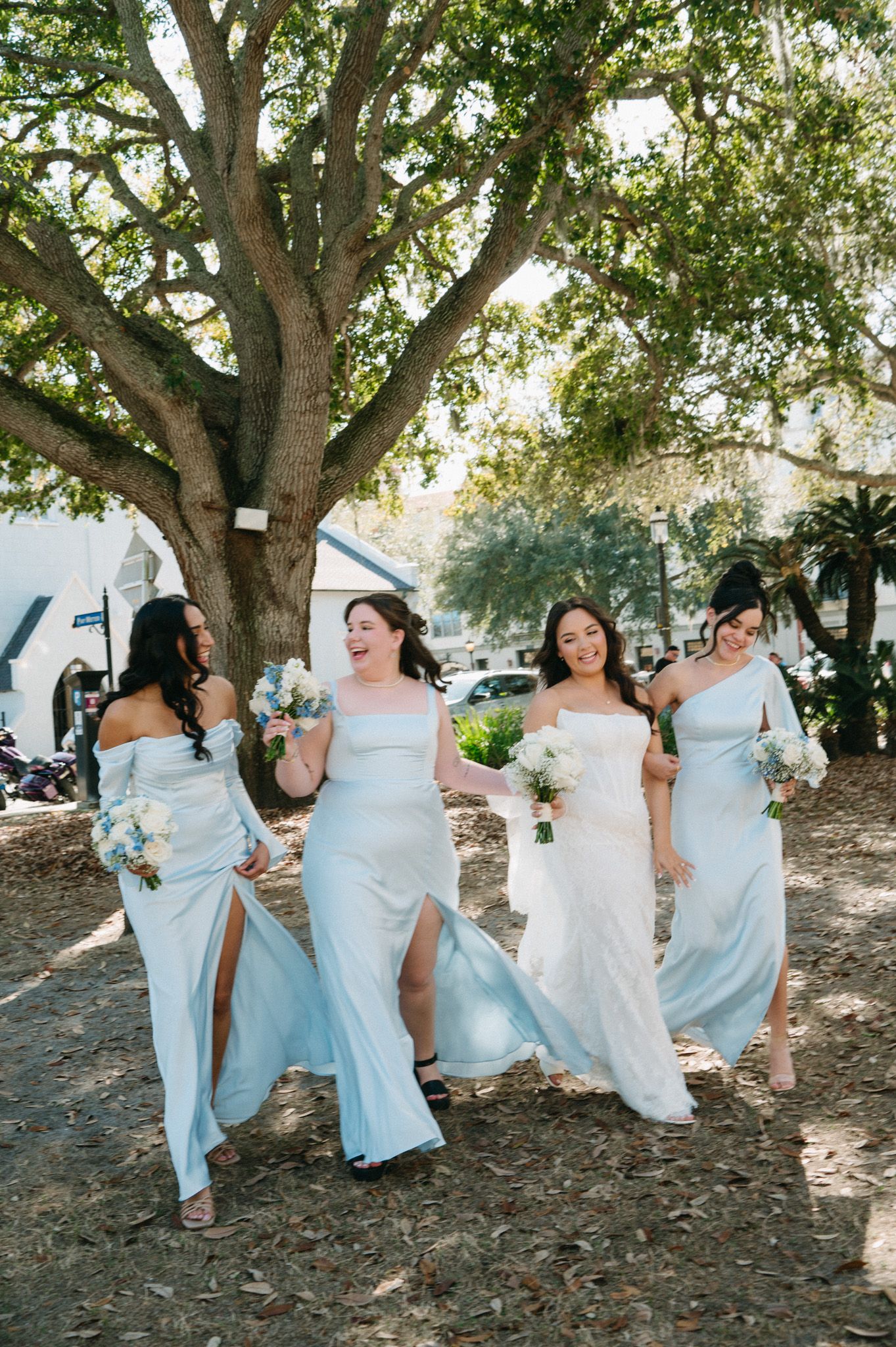 Bride and bridesmaids in light blue dresses walk joyfully under large oak tree on wedding day.
