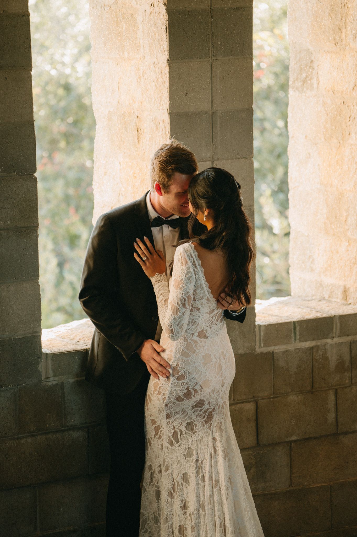 Bride in lace gown and groom in black tuxedo share romantic kiss by stone columns in soft natural light.
