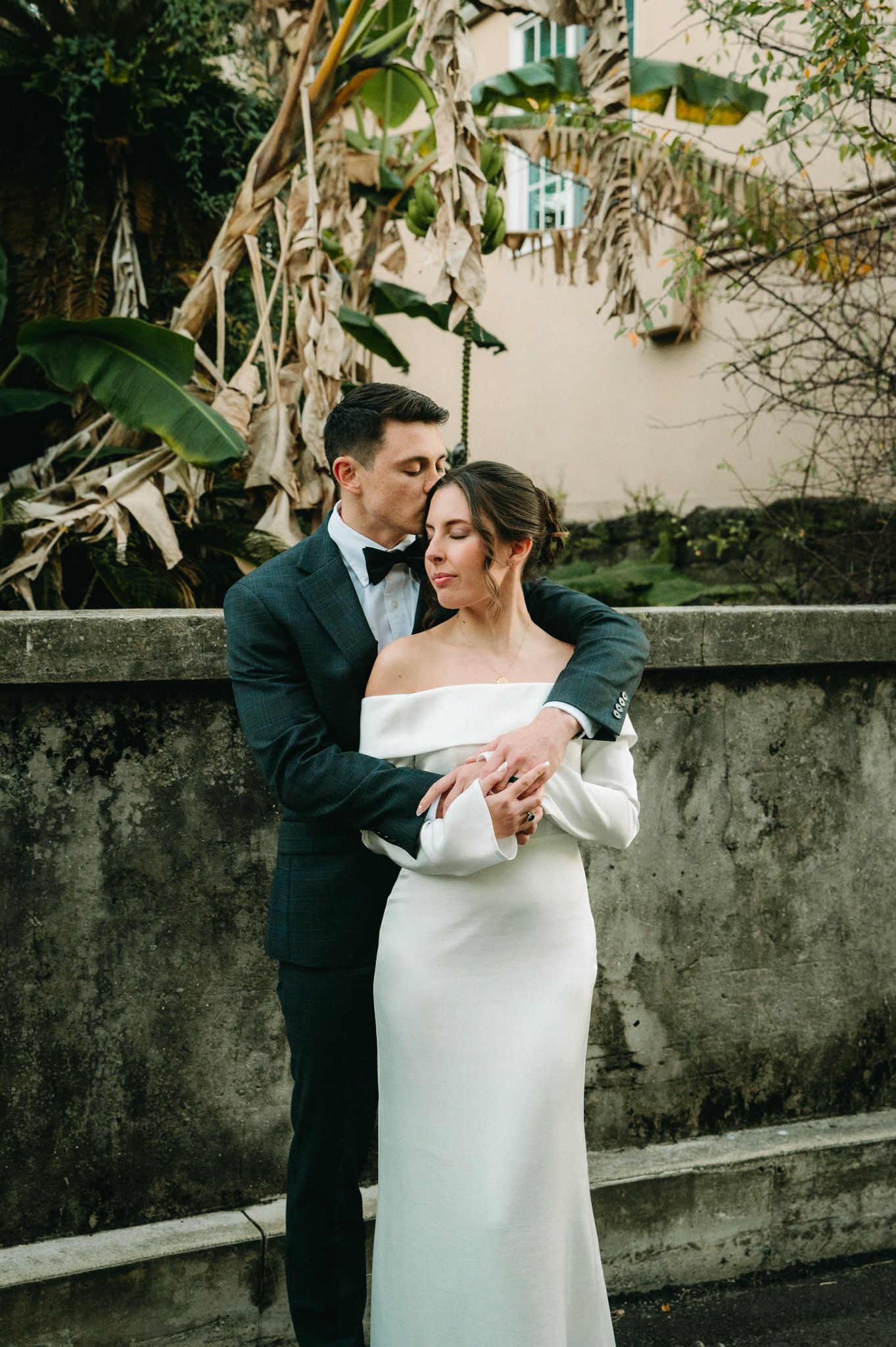 Groom in dark suit embraces bride in white off-shoulder wedding dress near stone wall with tropical plants.