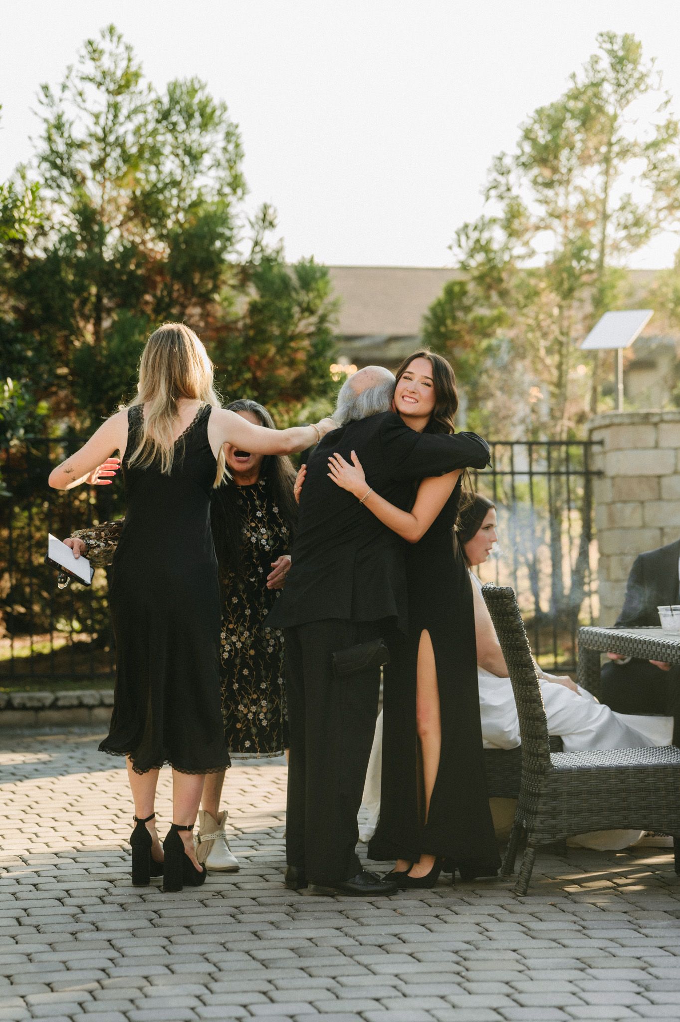 Bride in black slit dress joyfully embraces wedding guest on sunlit brick patio after outdoor ceremony.