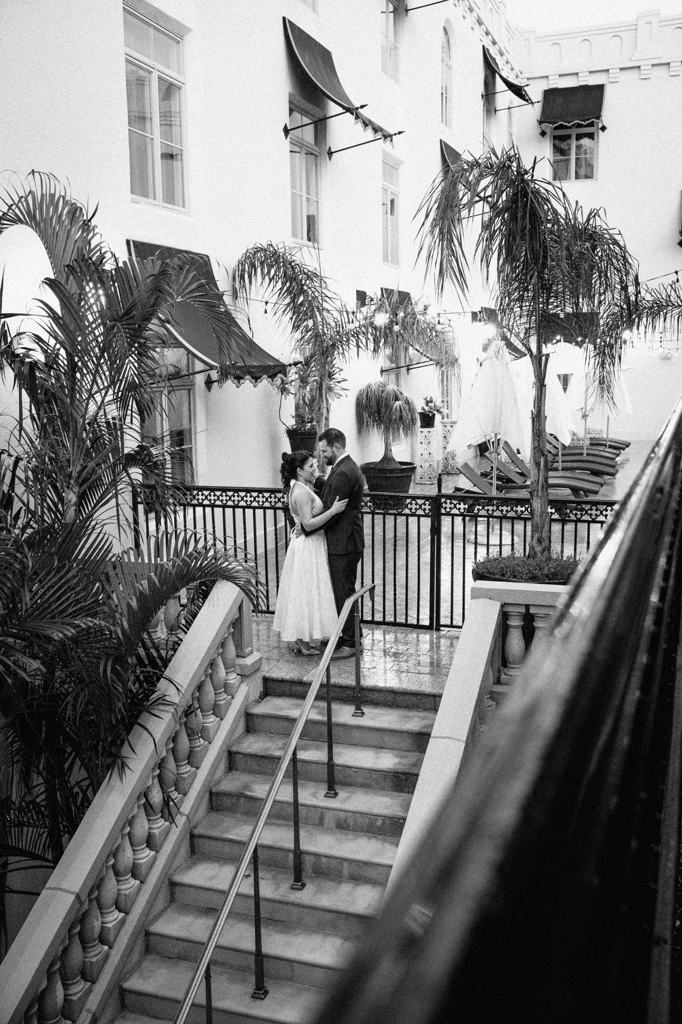 Bride and groom embracing on staircase of elegant hotel courtyard in black and white photo.