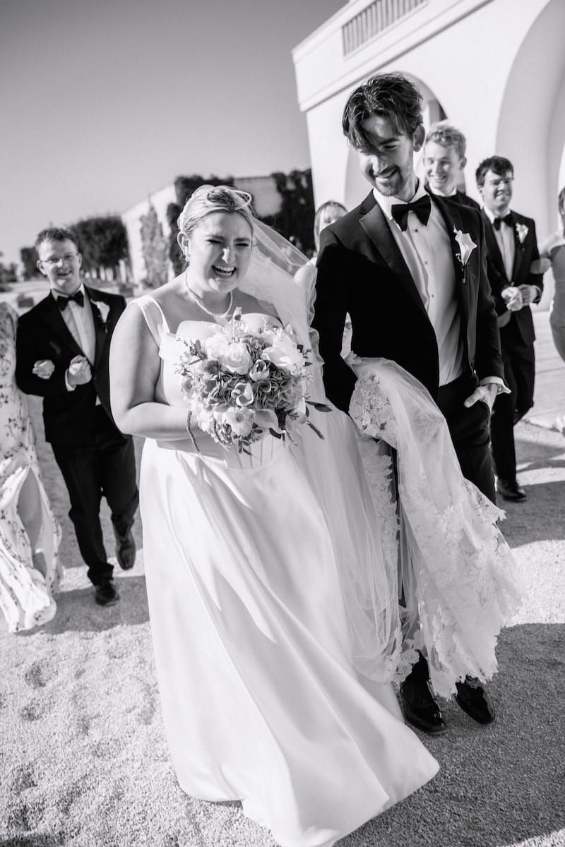 Black and white photo of a laughing bride holding bouquet walking with groom in tuxedo after wedding ceremony.