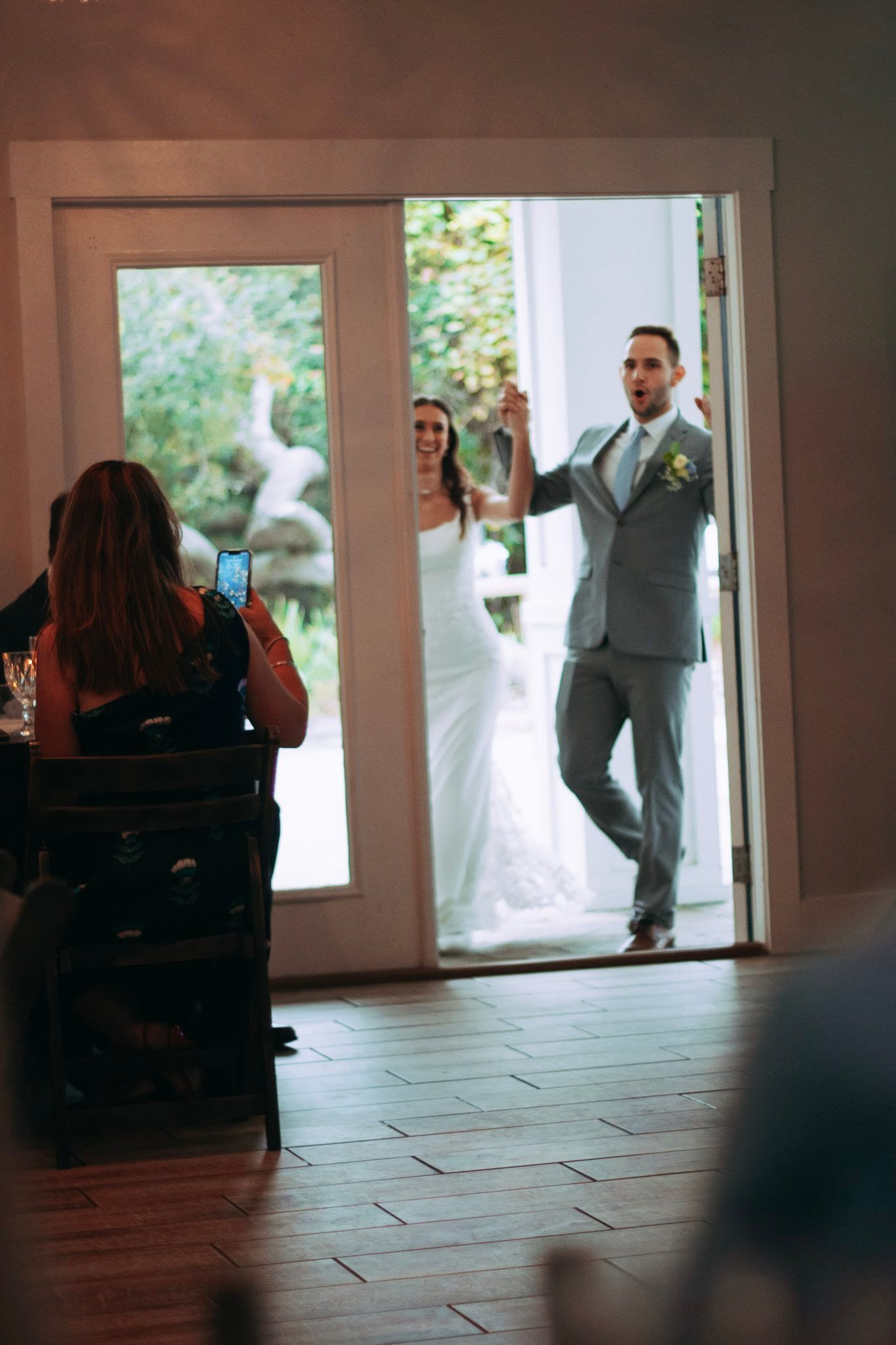 Bride and groom making grand entrance through doorway as guest captures moment on phone.