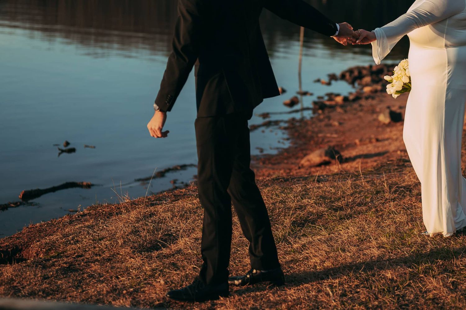 Groom holding the Bride's hand and looking back to her next to the lake bed with a warm and inviting style.