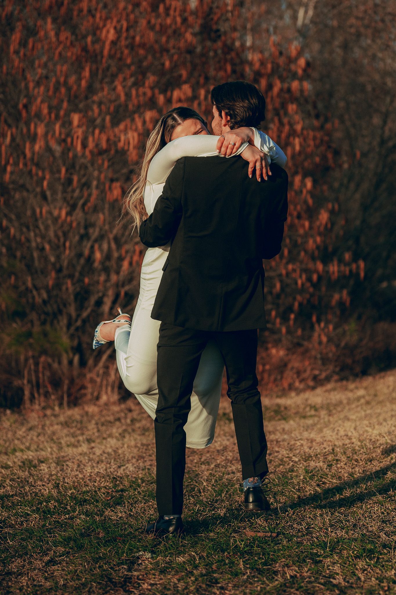 Groom in black suit lifting bride in white dress in romantic outdoor autumn setting.
