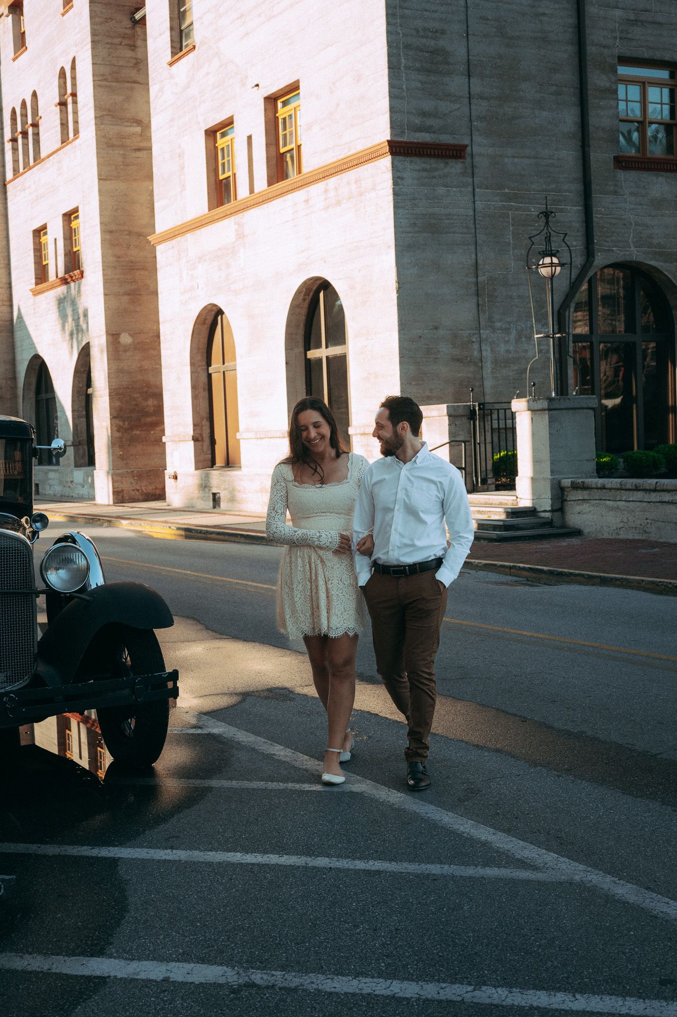 Engaged couple strolling and laughing on a sunny urban street lined with historic stone buildings.