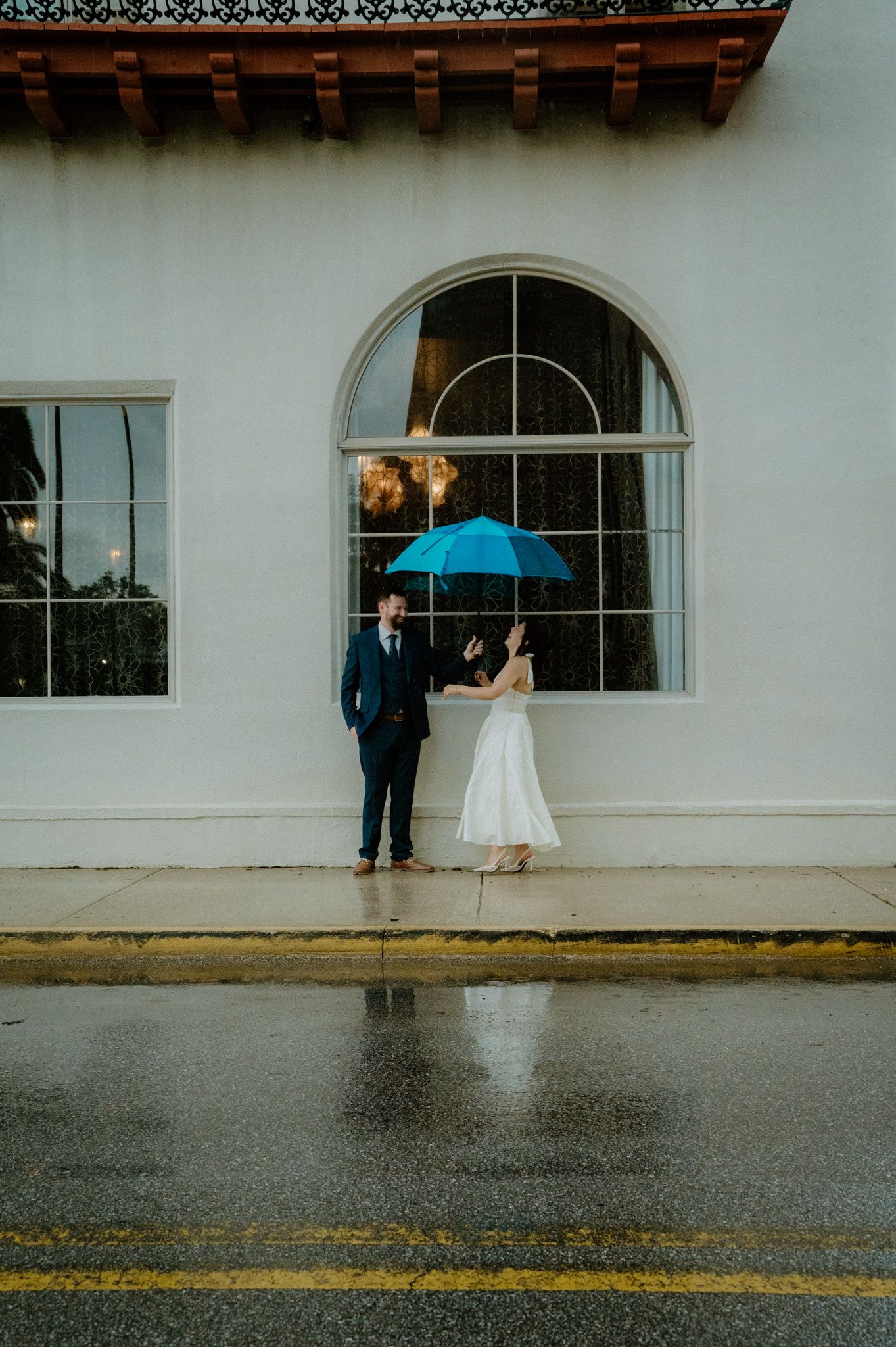 Bride with blue umbrella and groom stand outside white building on rainy wedding day.