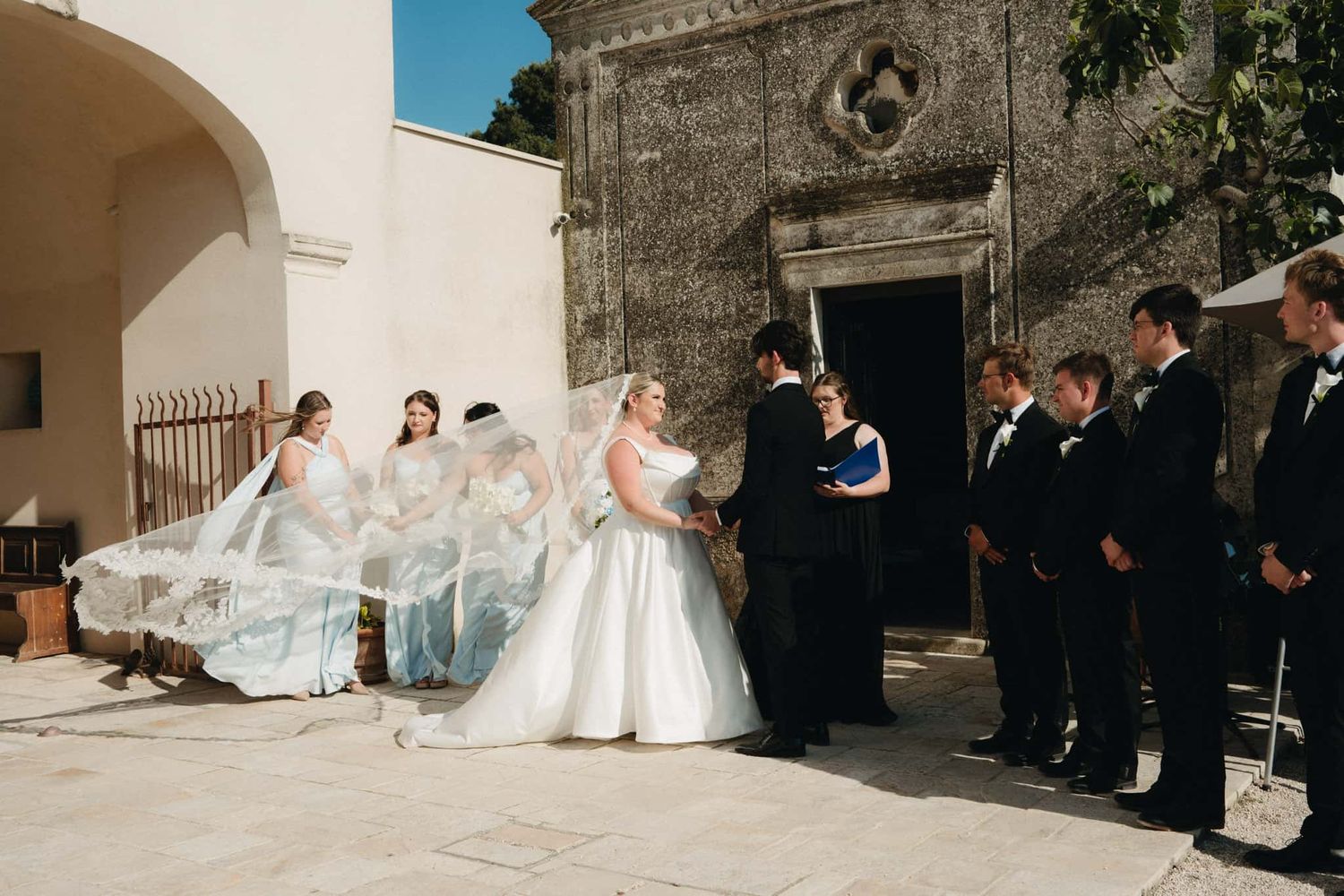 Bride in white ball gown with flowing veil held by bridesmaids during outdoor wedding ceremony at historic stone chapel.