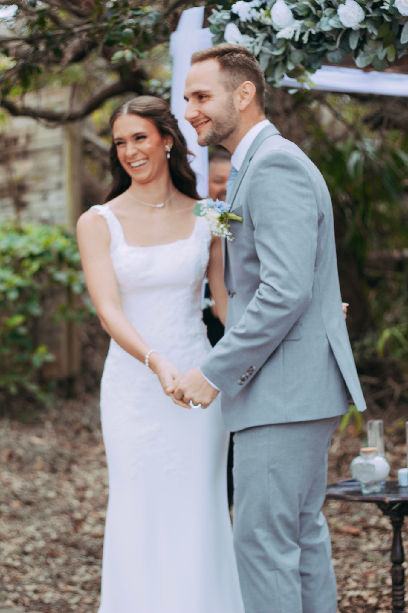 Bride and groom smile joyfully holding hands at outdoor garden wedding ceremony altar.