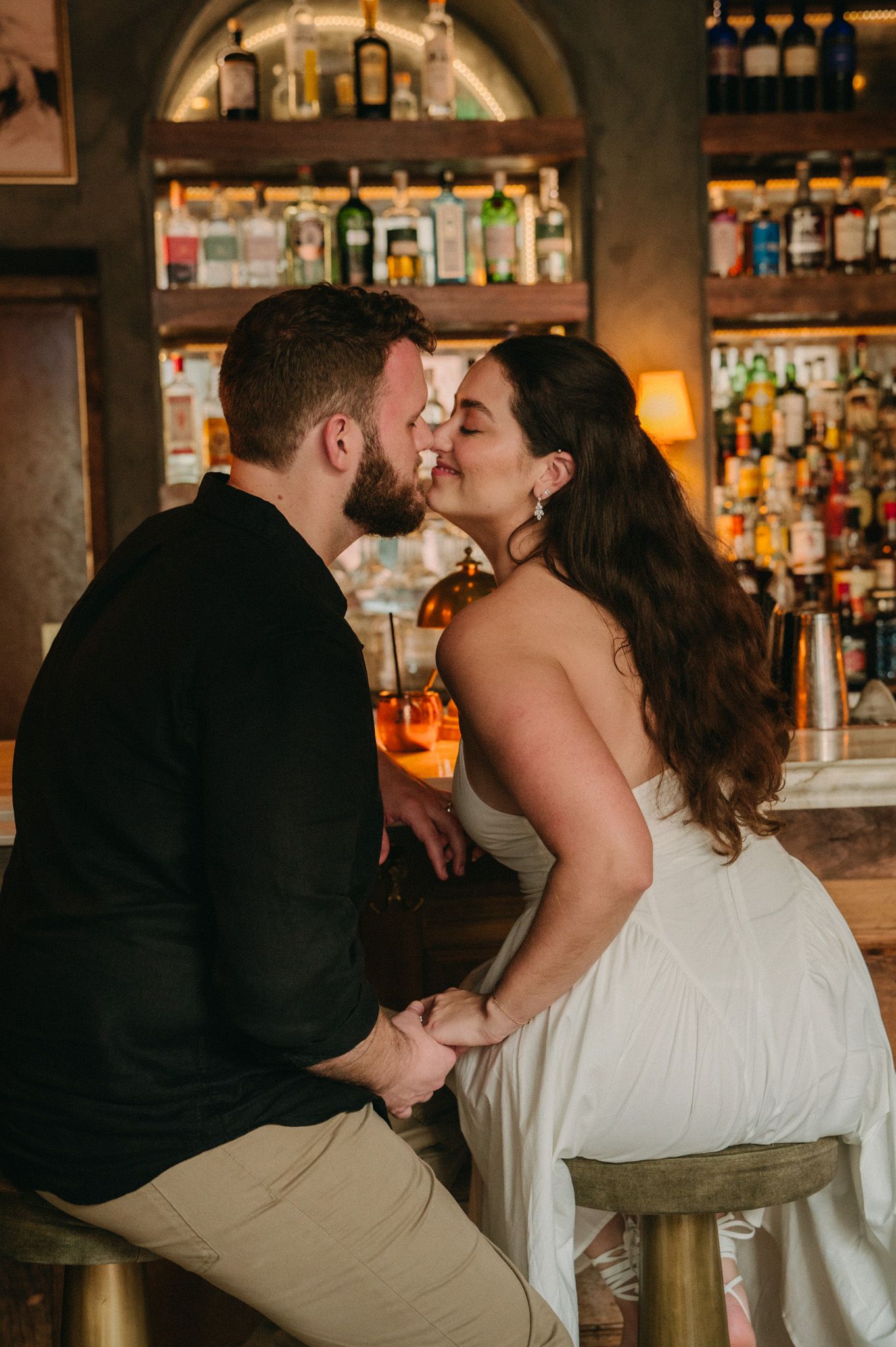 Couple kissing at a warmly lit bar while holding hands on bar stools.