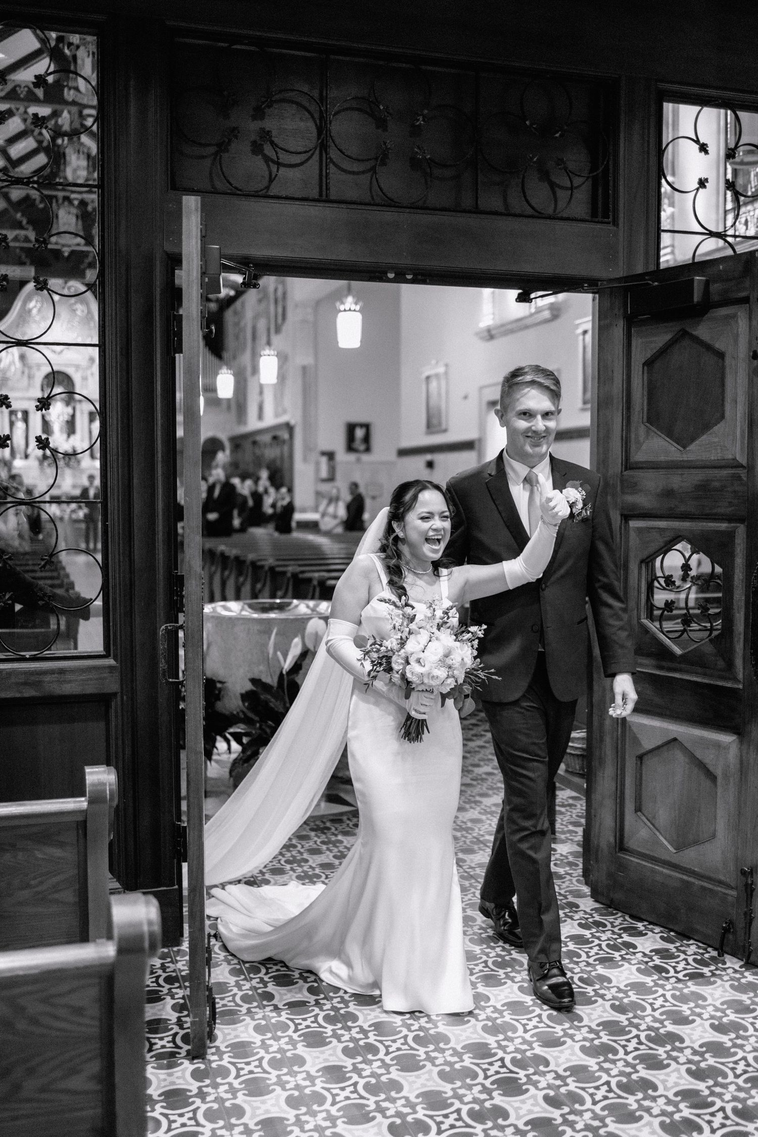 Black and white photo of a joyful bride and groom exiting through ornate wooden doors after their wedding ceremony.
