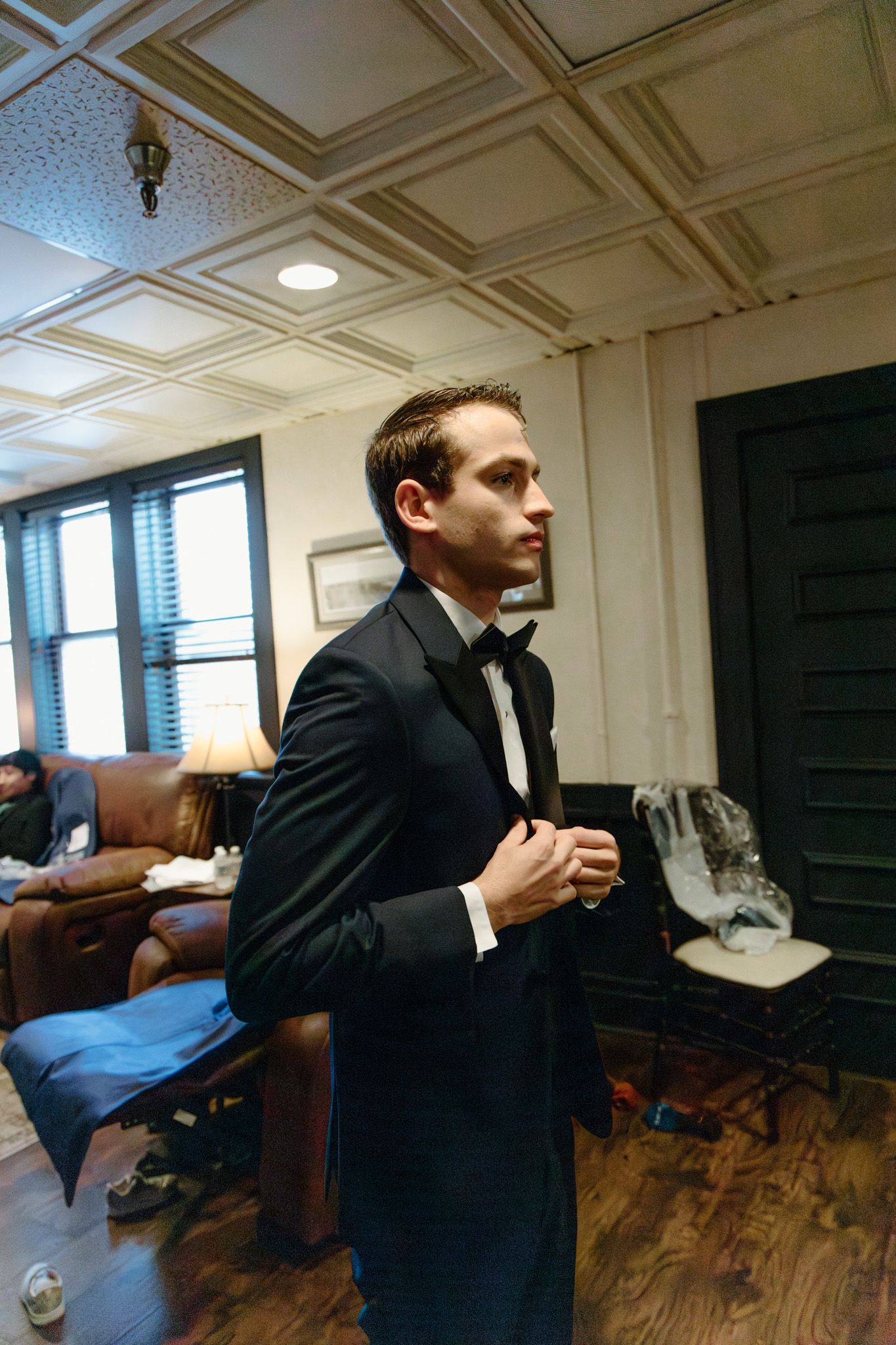 A young man in a black tuxedo adjusts his jacket in an elegantly decorated room with coffered ceilings.