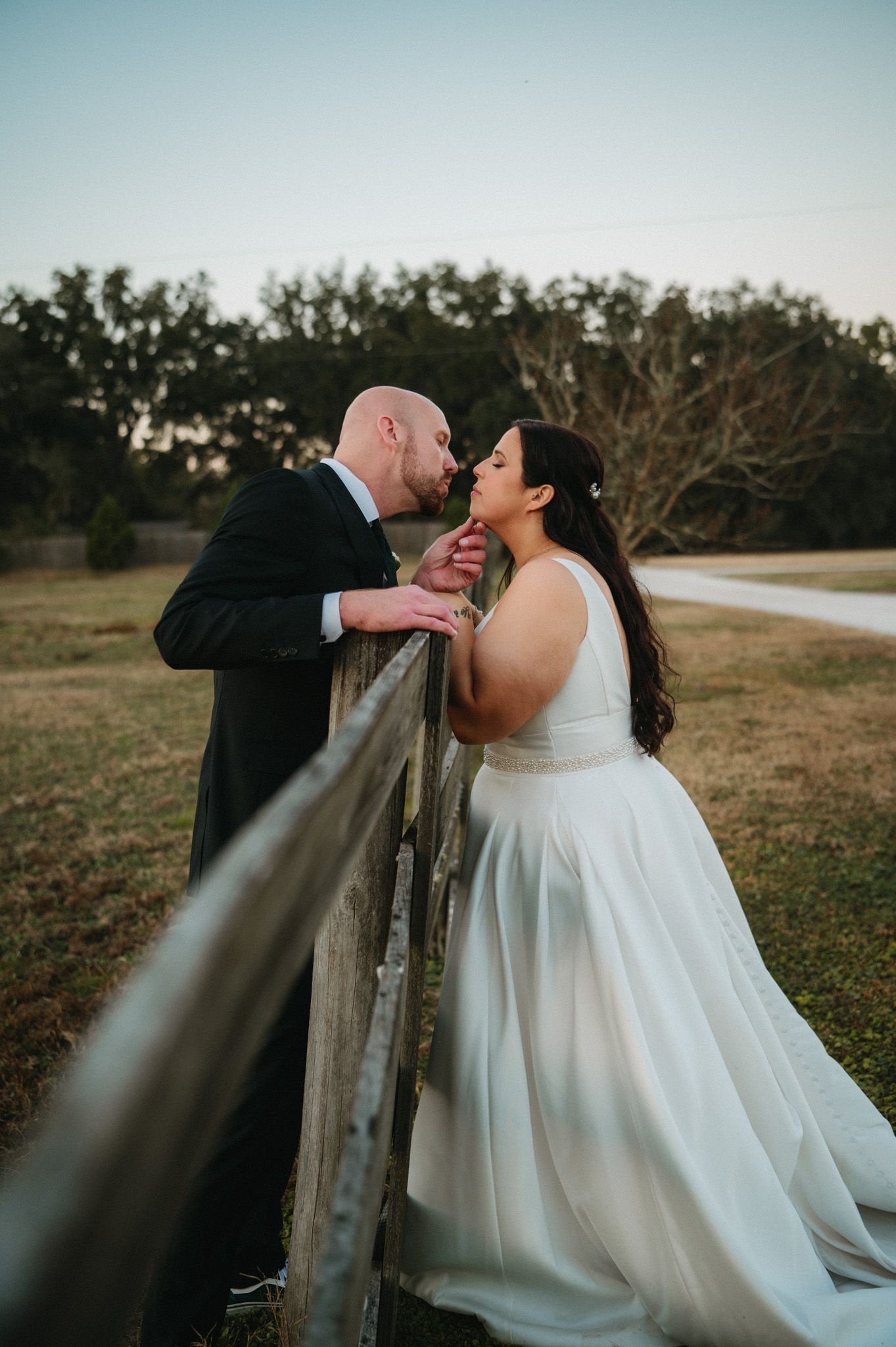 Bride and groom sharing a tender kiss over a rustic wooden fence at sunset in an open field.