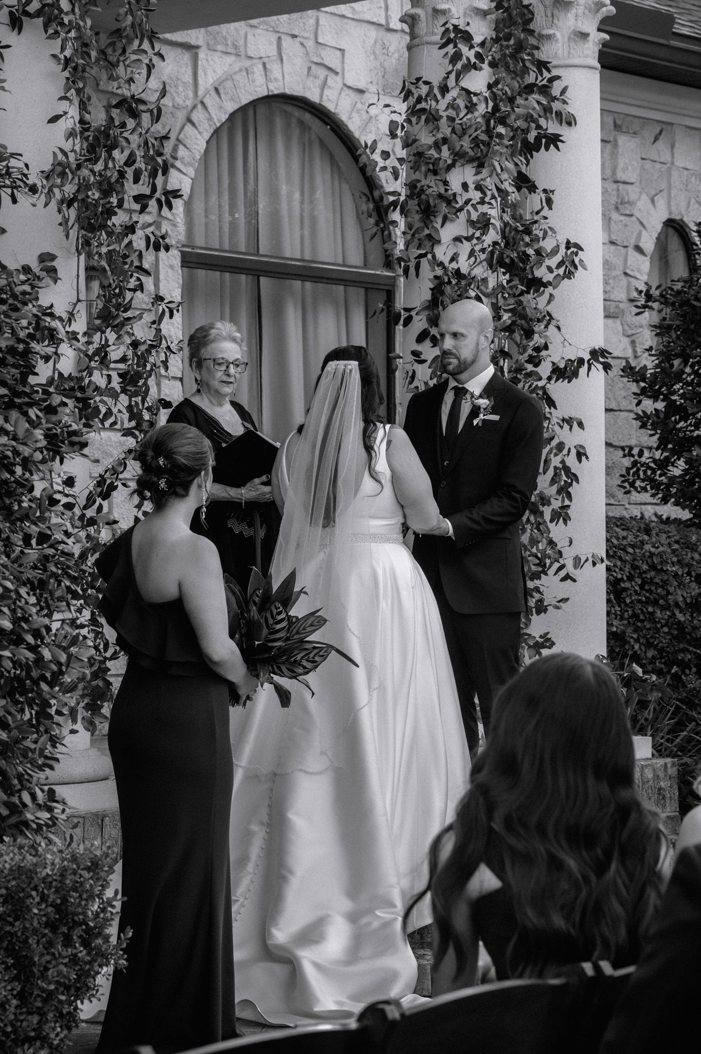Bride and groom exchange vows before officiant at elegant stone chapel entrance with ivy in black and white.