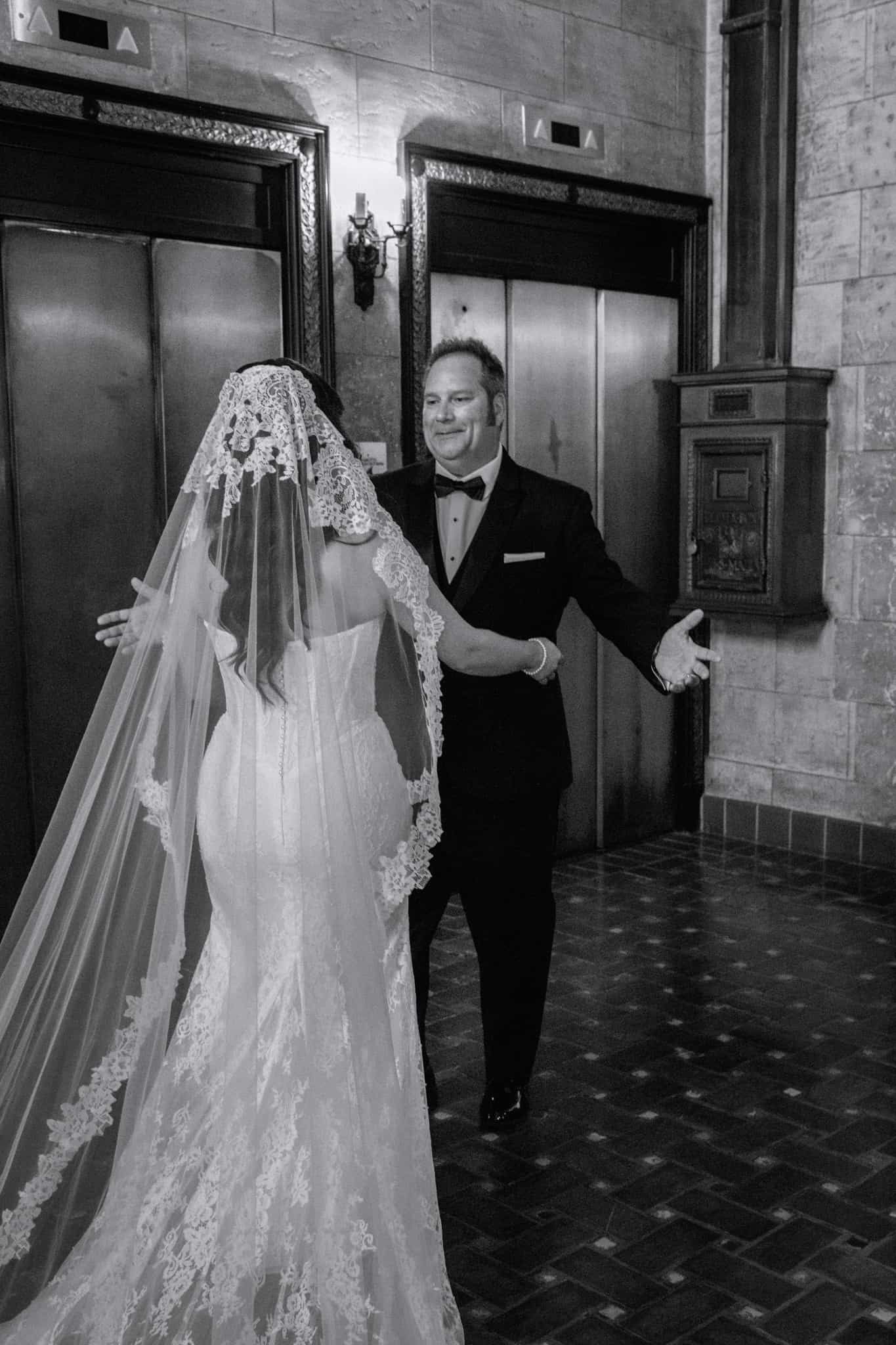 Black and white photo of a bride in lace wedding gown and veil sharing a first look moment with dad in tuxedo at The Treasury on the Plaza.