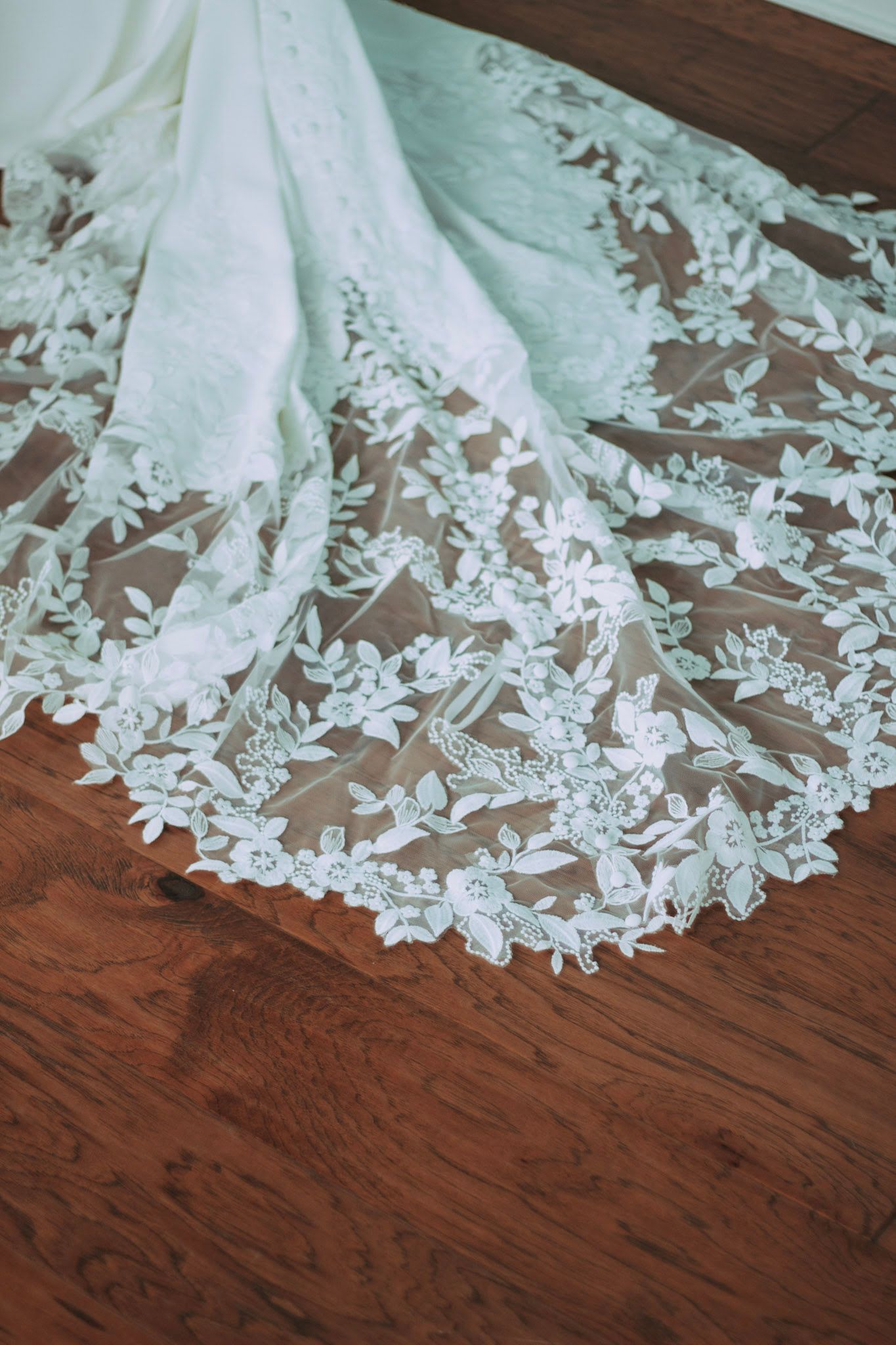 Intricate floral lace detail on white wedding dress train spread on hardwood floor.