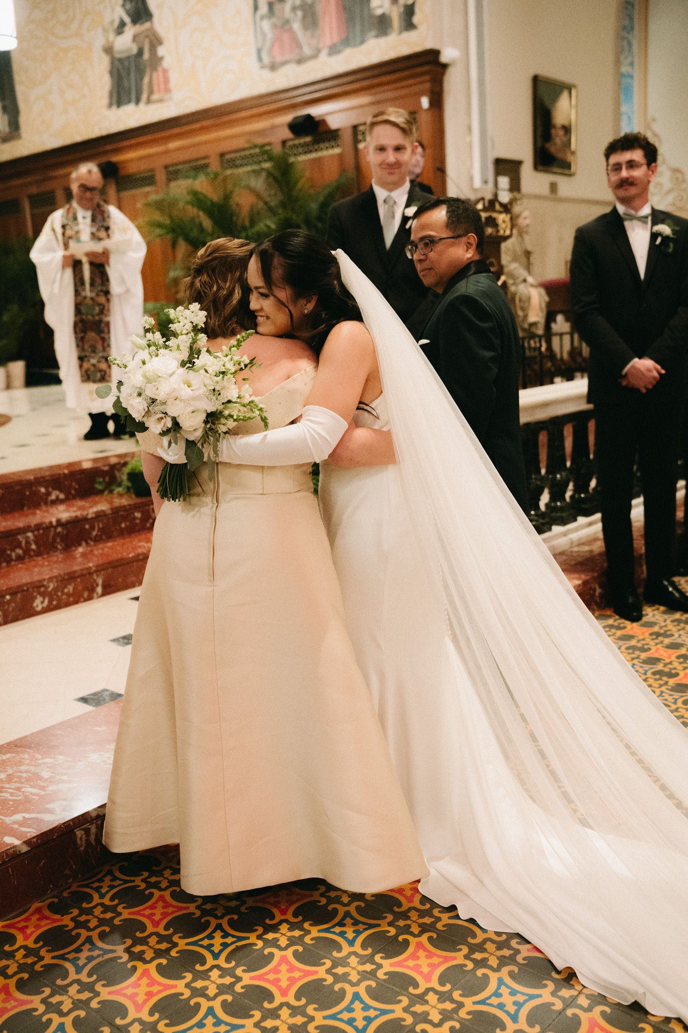 Two brides share a heartfelt hug at the altar during their church wedding ceremony, one holding a white floral bouquet.