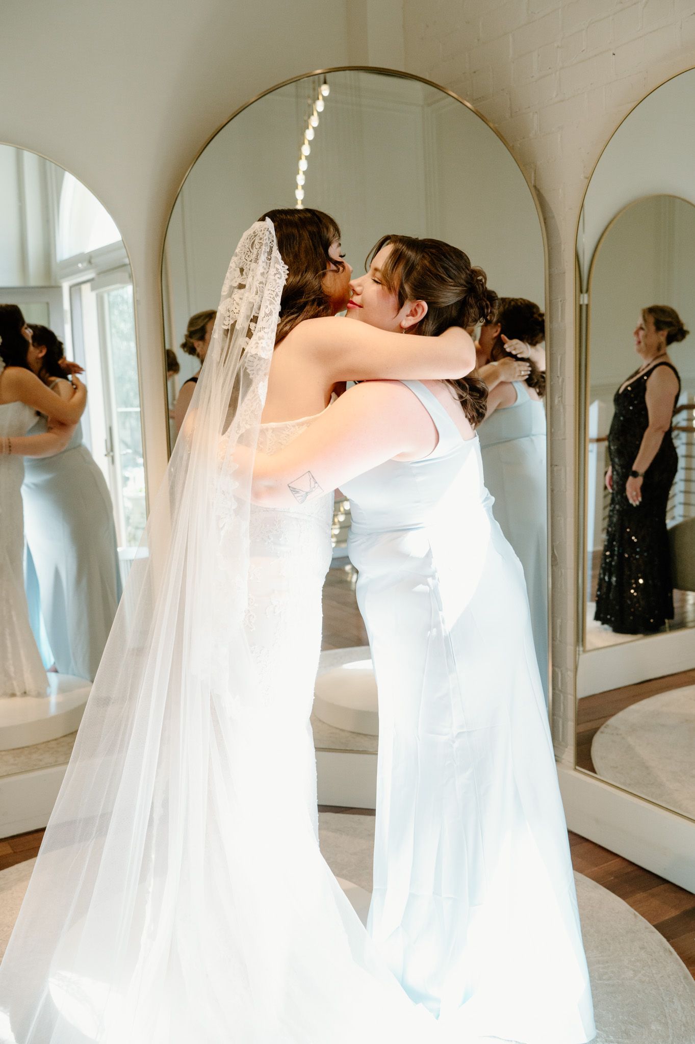 Bride in white gown and veil embracing bridesmaid in light blue dress near arched mirrors.