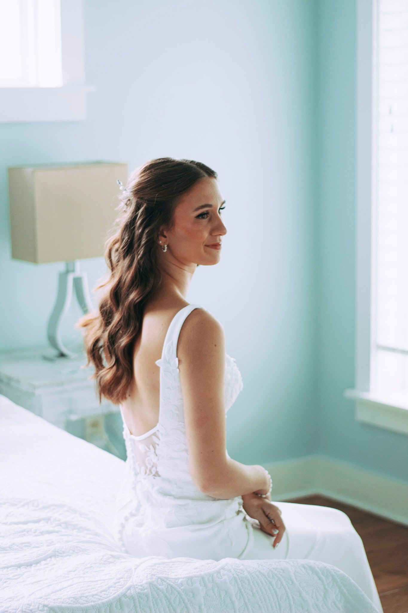 Bride in white lace wedding dress sitting on bed in teal bedroom smiling.