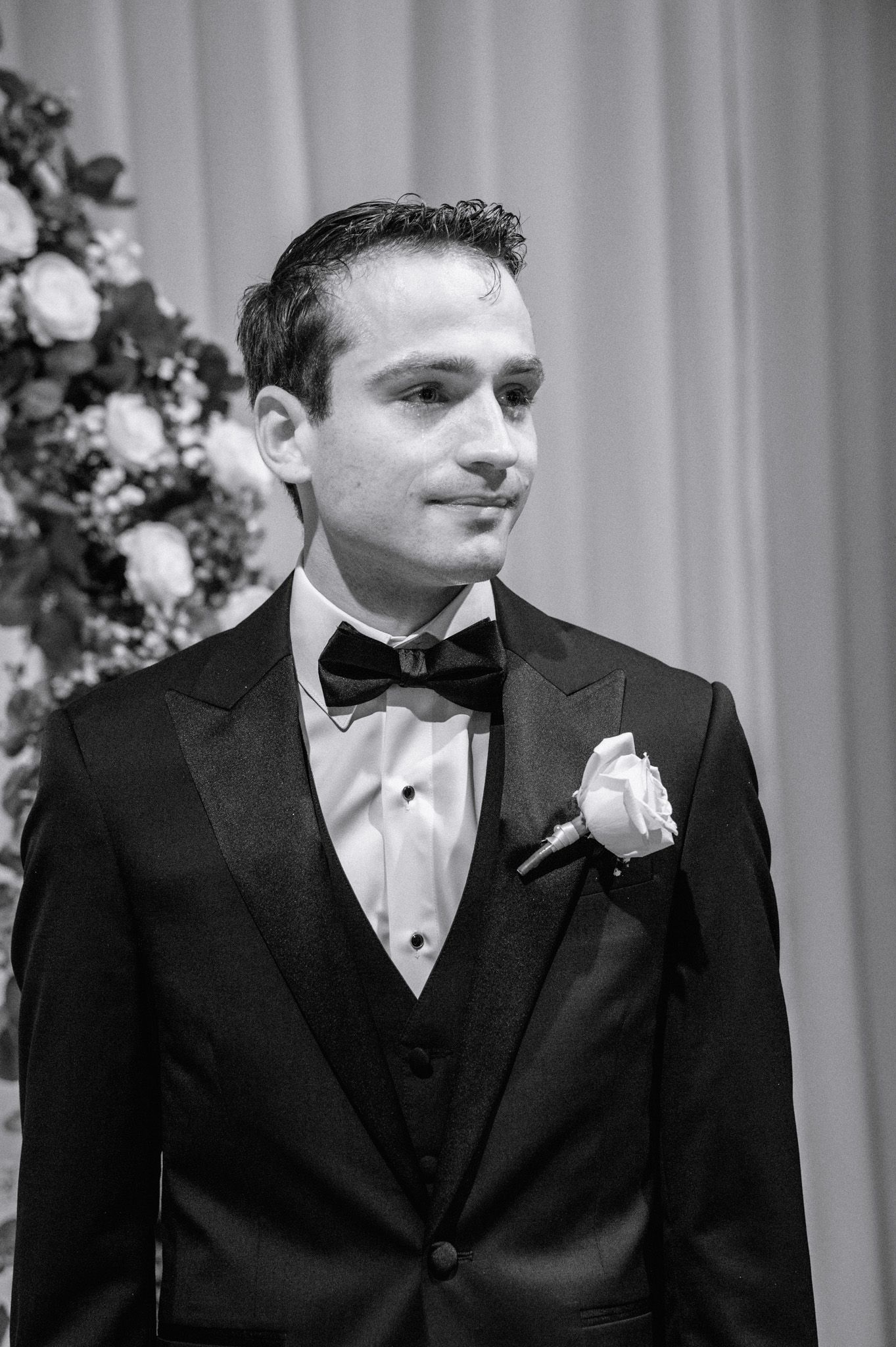 Black and white portrait of groom in tuxedo with rose boutonniere standing at wedding altar.