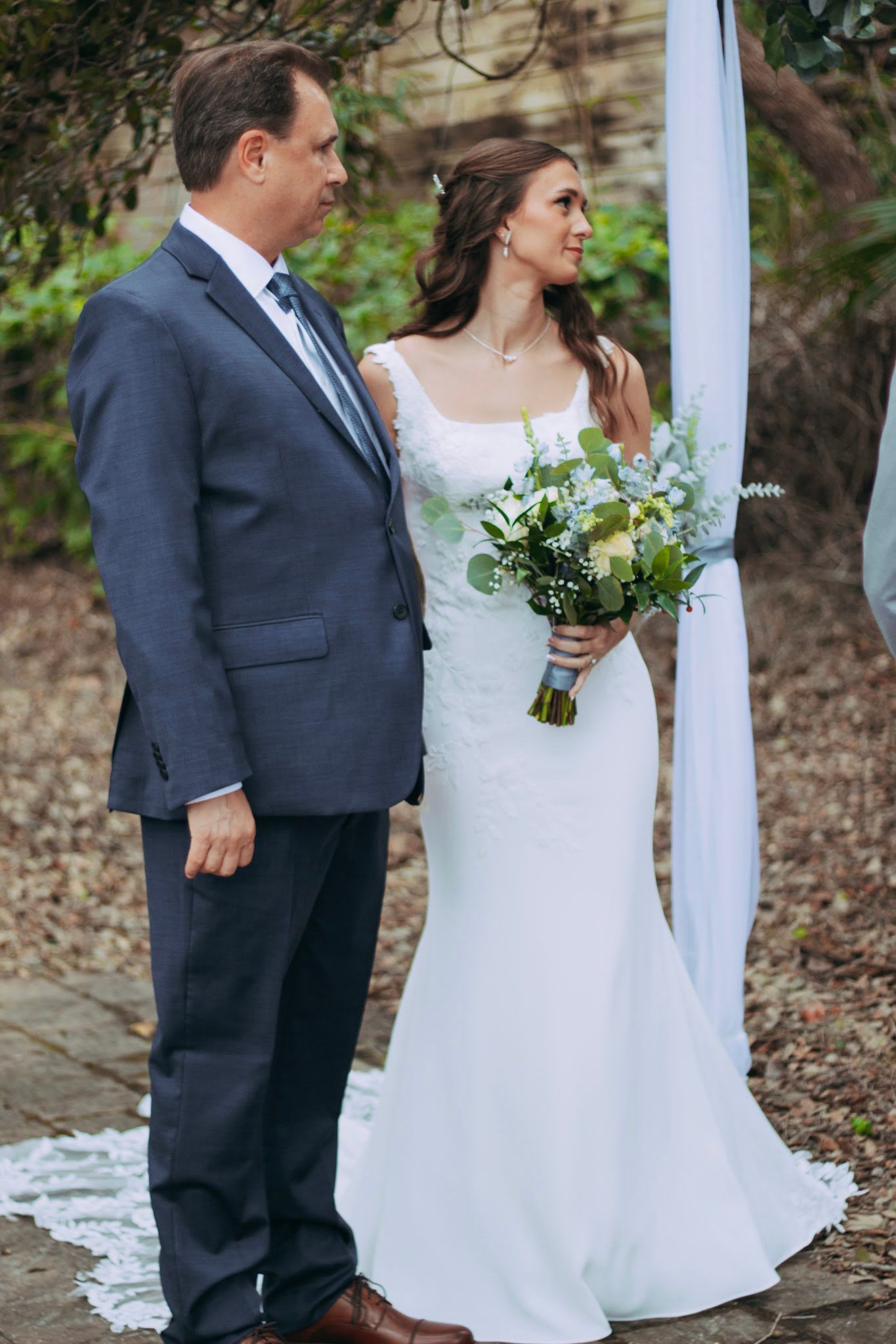 Bride holding white bouquet stands with escort beside floral arch at outdoor wedding ceremony.