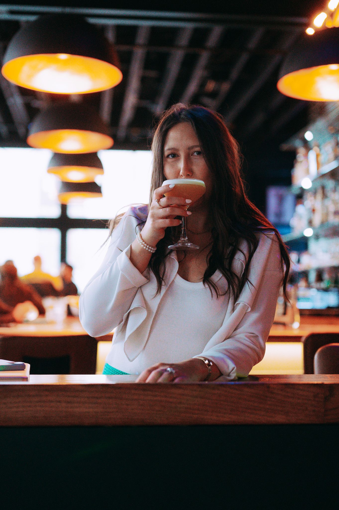 Woman sipping a cocktail at a dimly lit bar with warm pendant lights overhead.