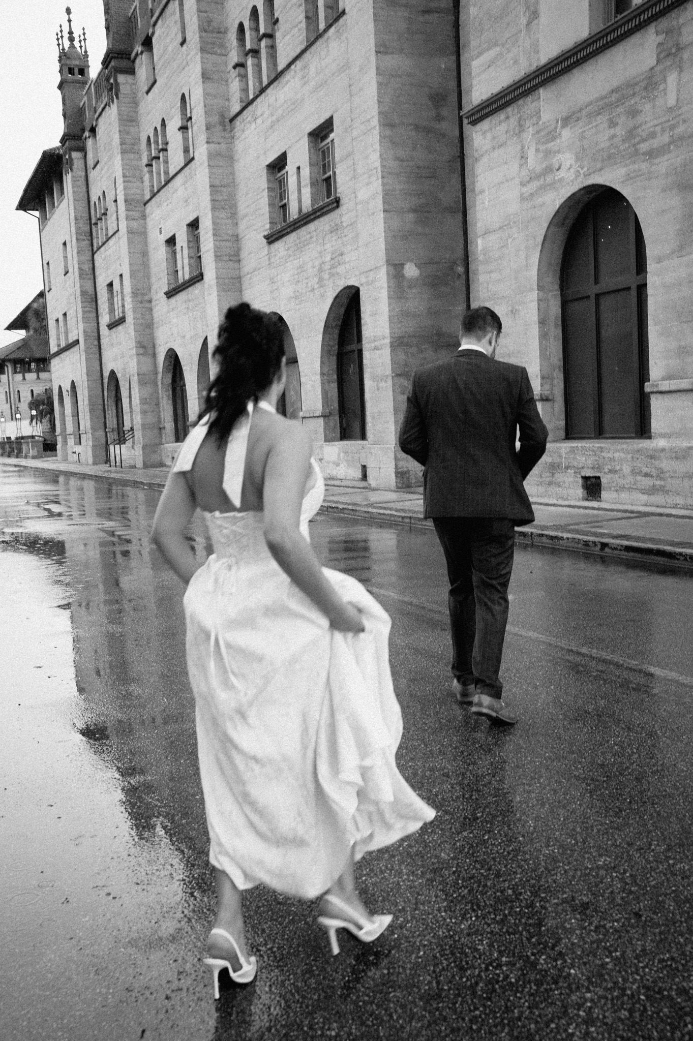 Black and white photo of bride and groom walking in rain along a historic European building.