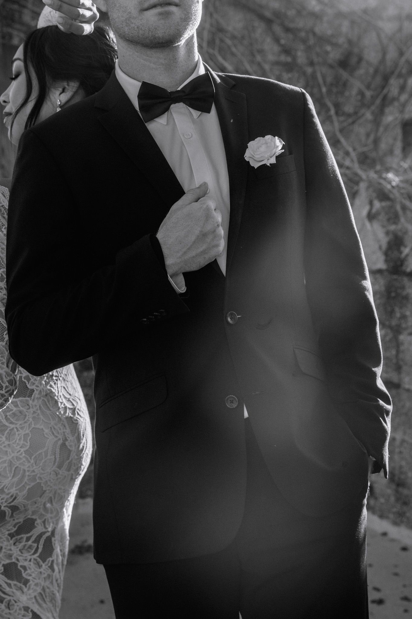 Black and white close-up of groom in tuxedo with bow tie and white rose boutonniere at wedding.