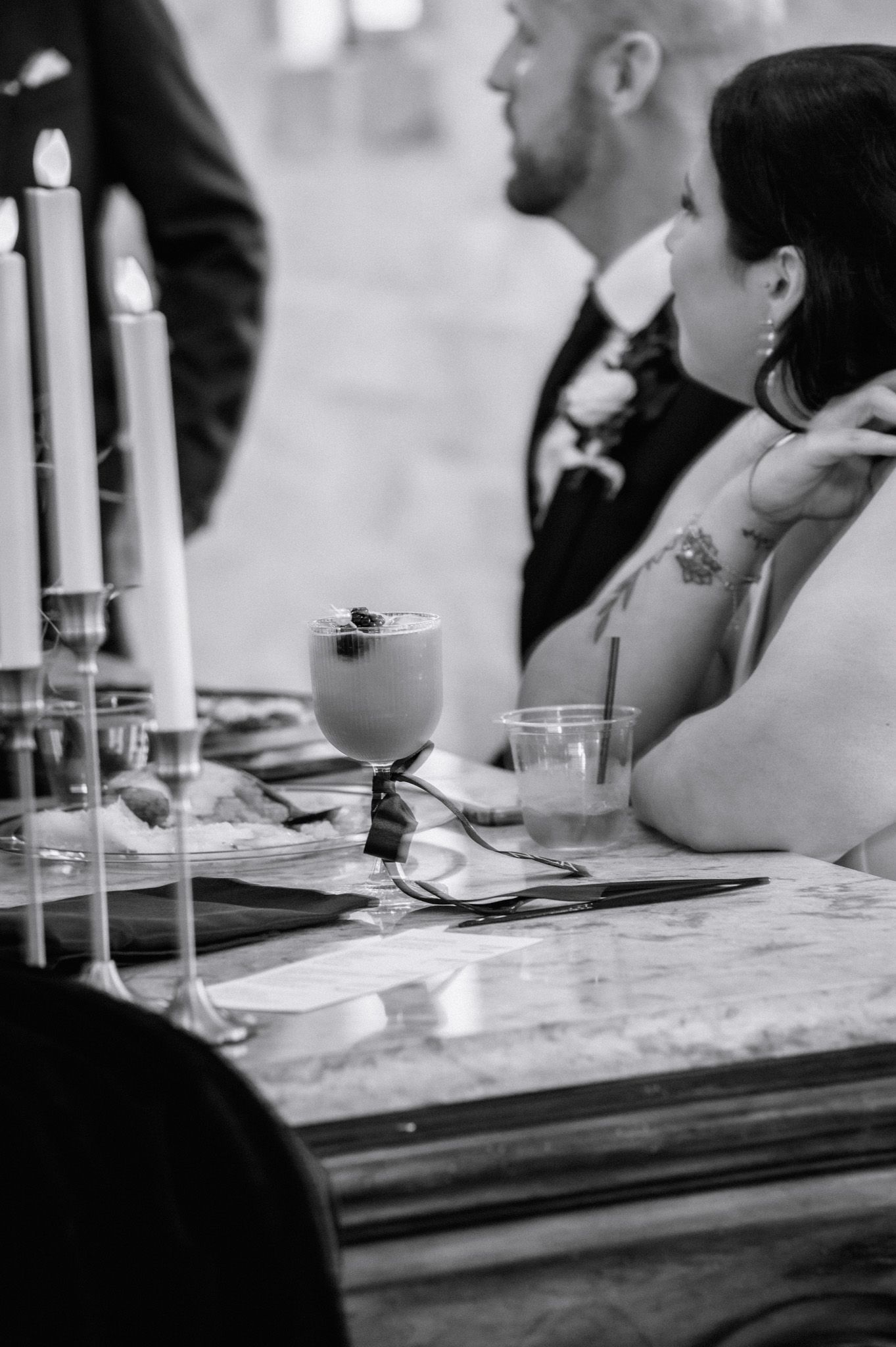 Bride with tattoos sits at marble sweetheart table with elegant cocktail and candles at wedding reception.