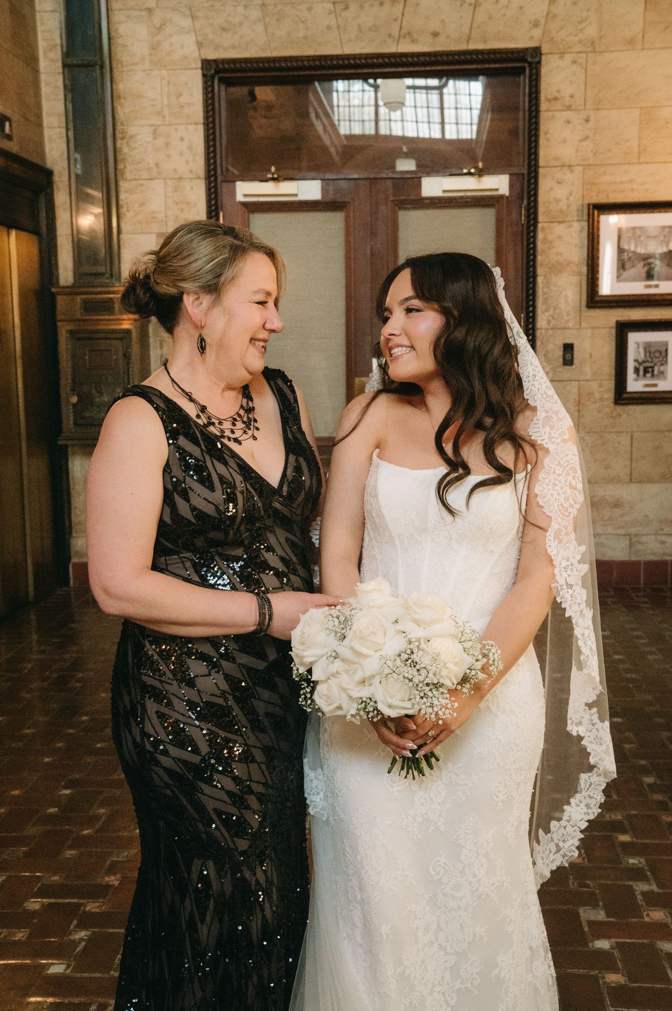 Bride in lace gown holding white bouquet stands with woman in black sequin dress in elegant venue.