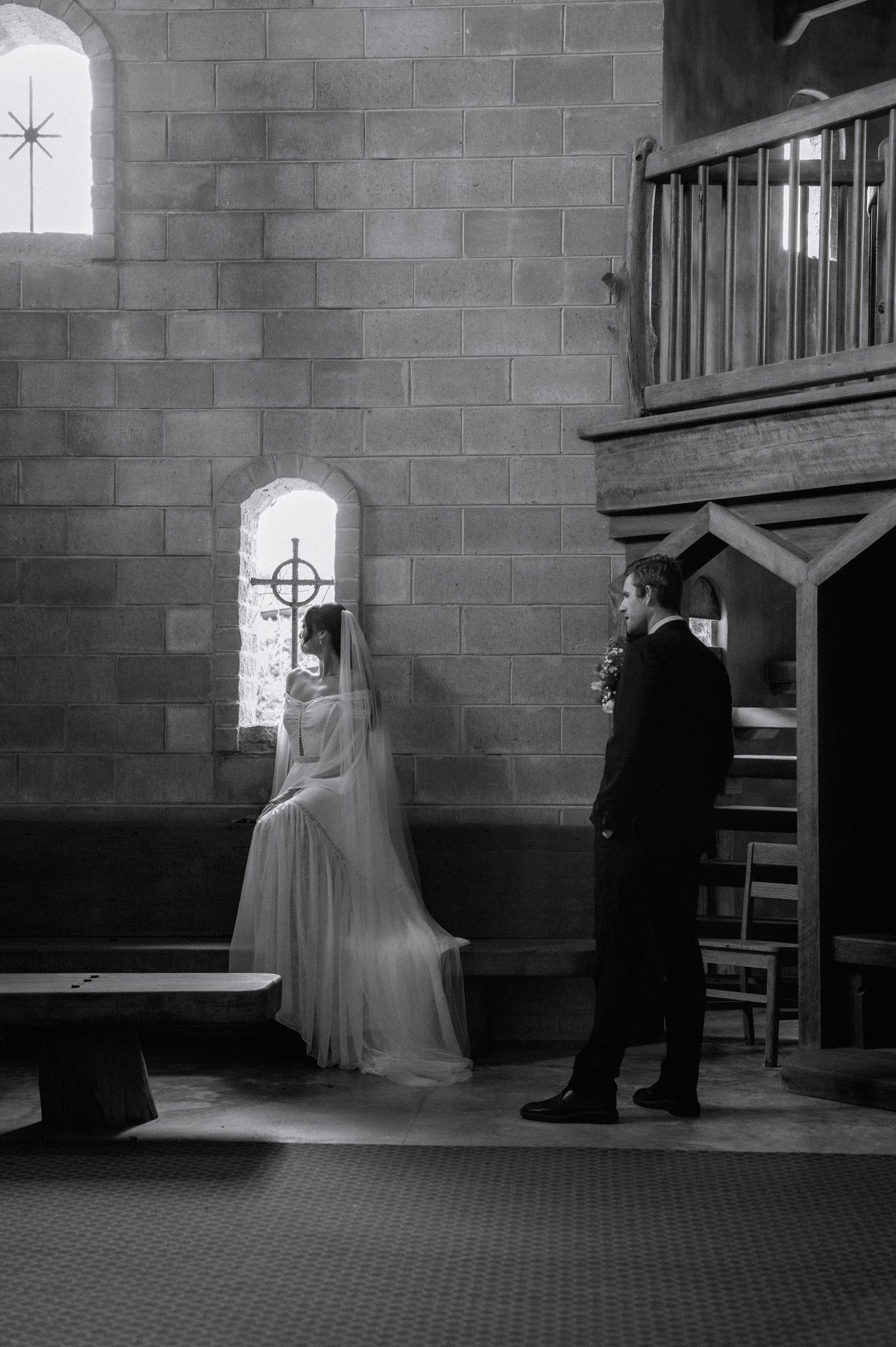 Black and white photo of bride in veil seated by arched church window as groom stands nearby in stone chapel.