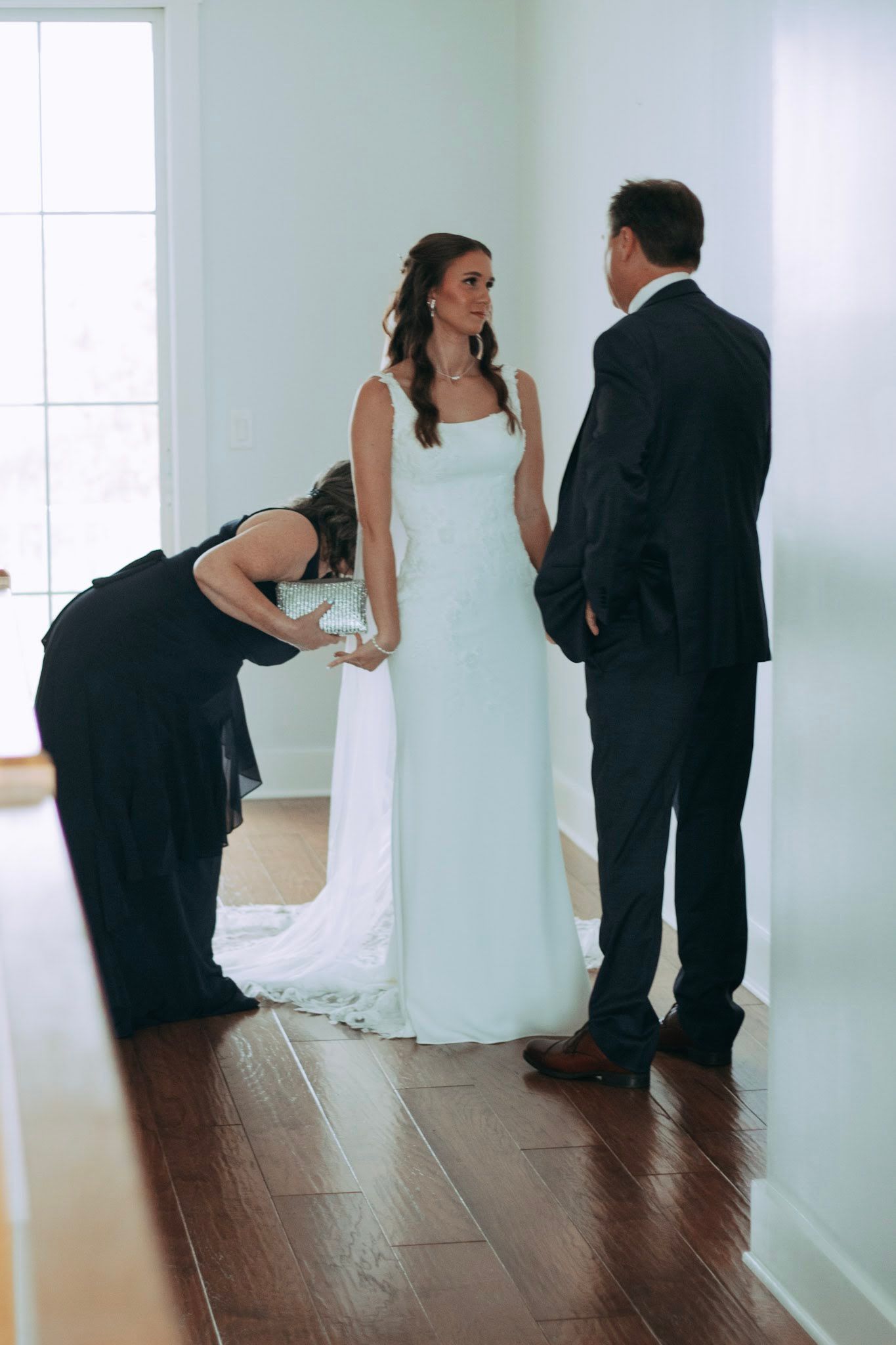 Bride in white gown speaking with father in black suit while bridesmaid adjusts her dress.